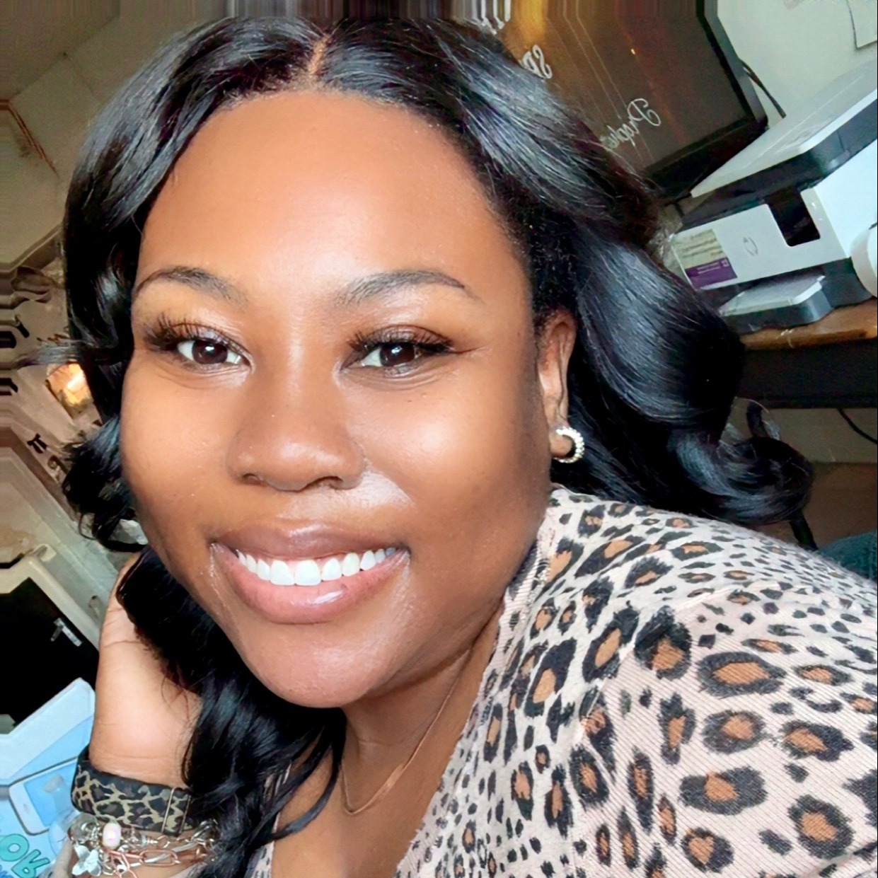 A woman with long, curly black hair wearing a leopard print top. She is smiling and has her hand on her cheek.