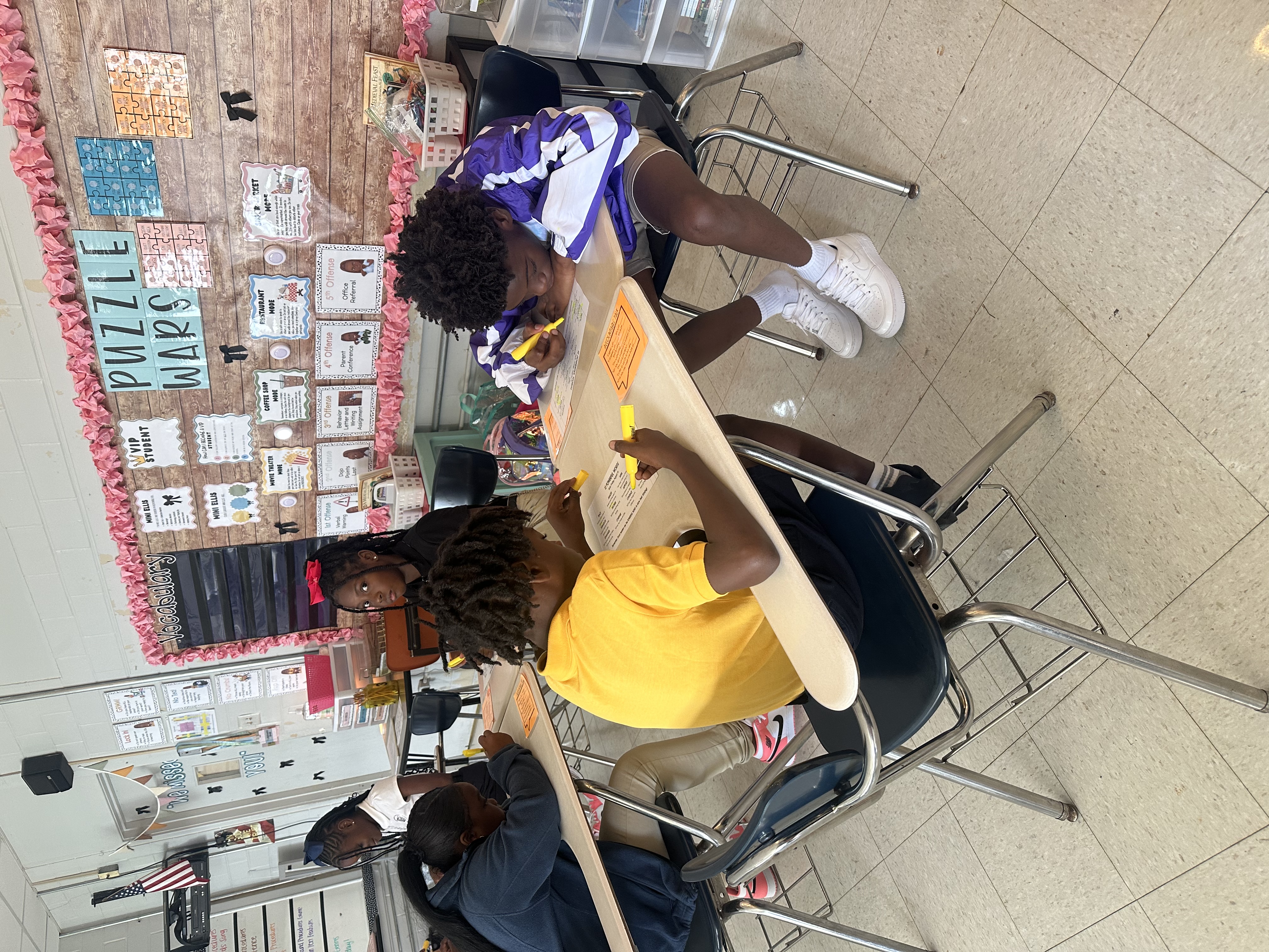 Four students seated at desks in a classroom, working on papers and using pens. The classroom features a bulletin board.