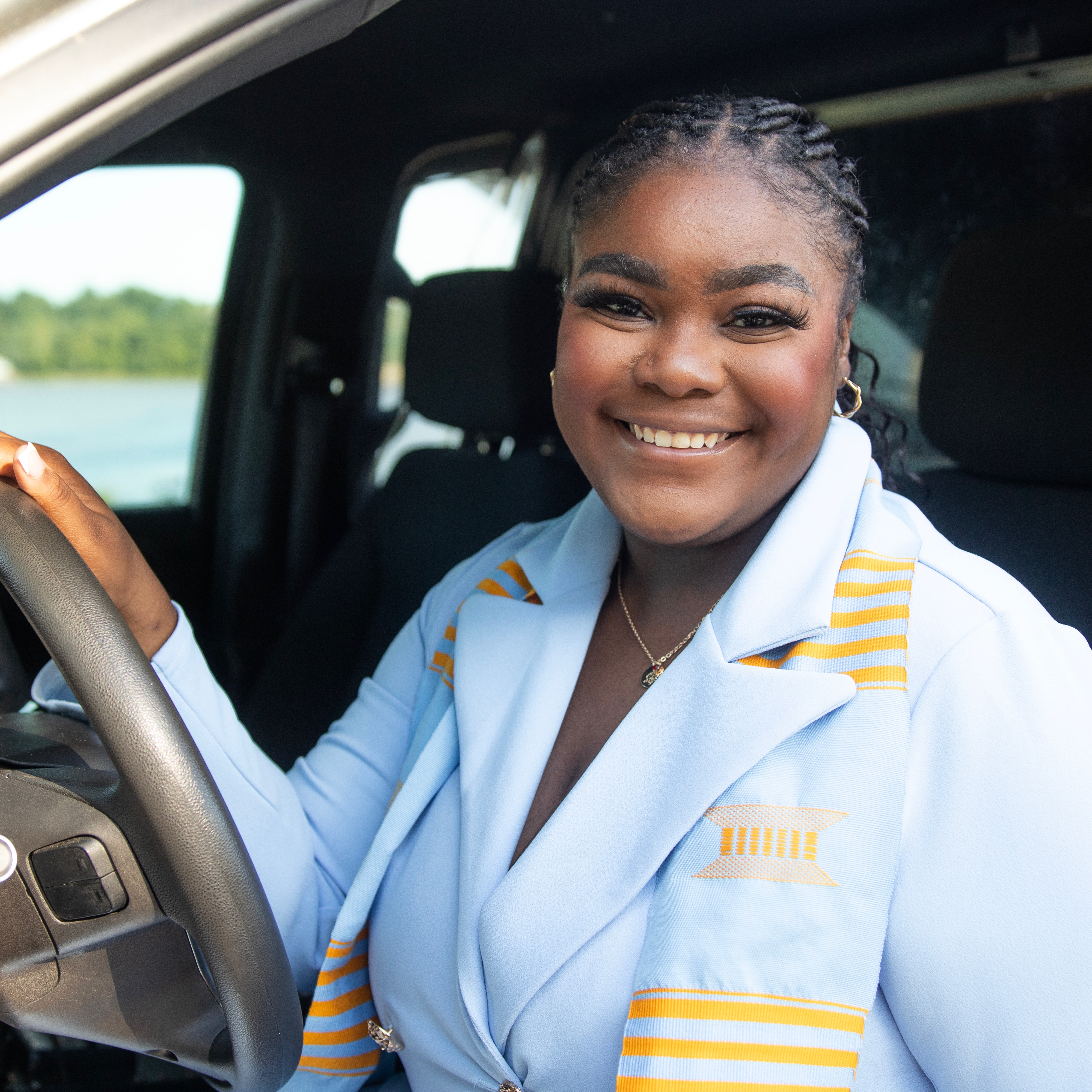 Person in a light blue suit sitting in a car and smiling, holding the steering wheel.