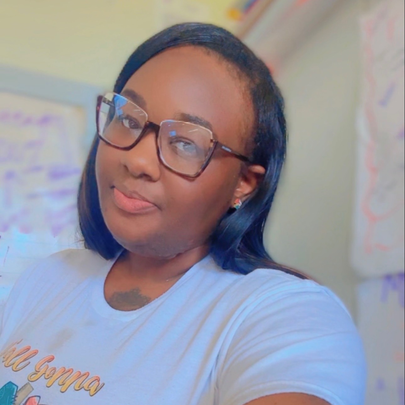 Person with glasses and a white t-shirt with colorful writing. In a room with white walls and papers.