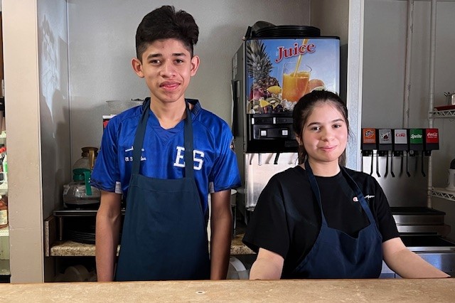a teenage  boy and girl wearing aprons behind restaurant counter