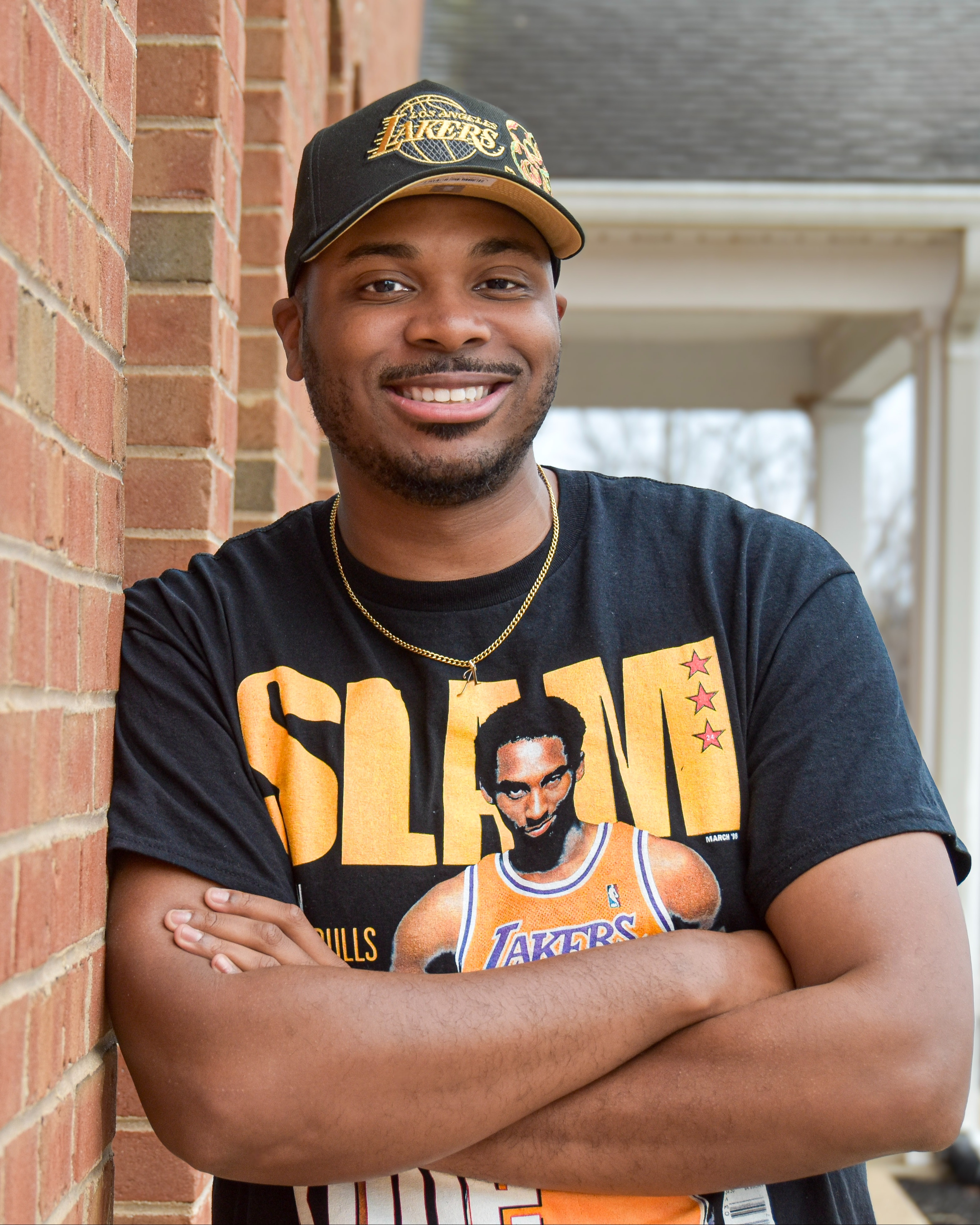 A man leans against a brick wall with arms crossed. He wears a Lakers t-shirt and a black cap.