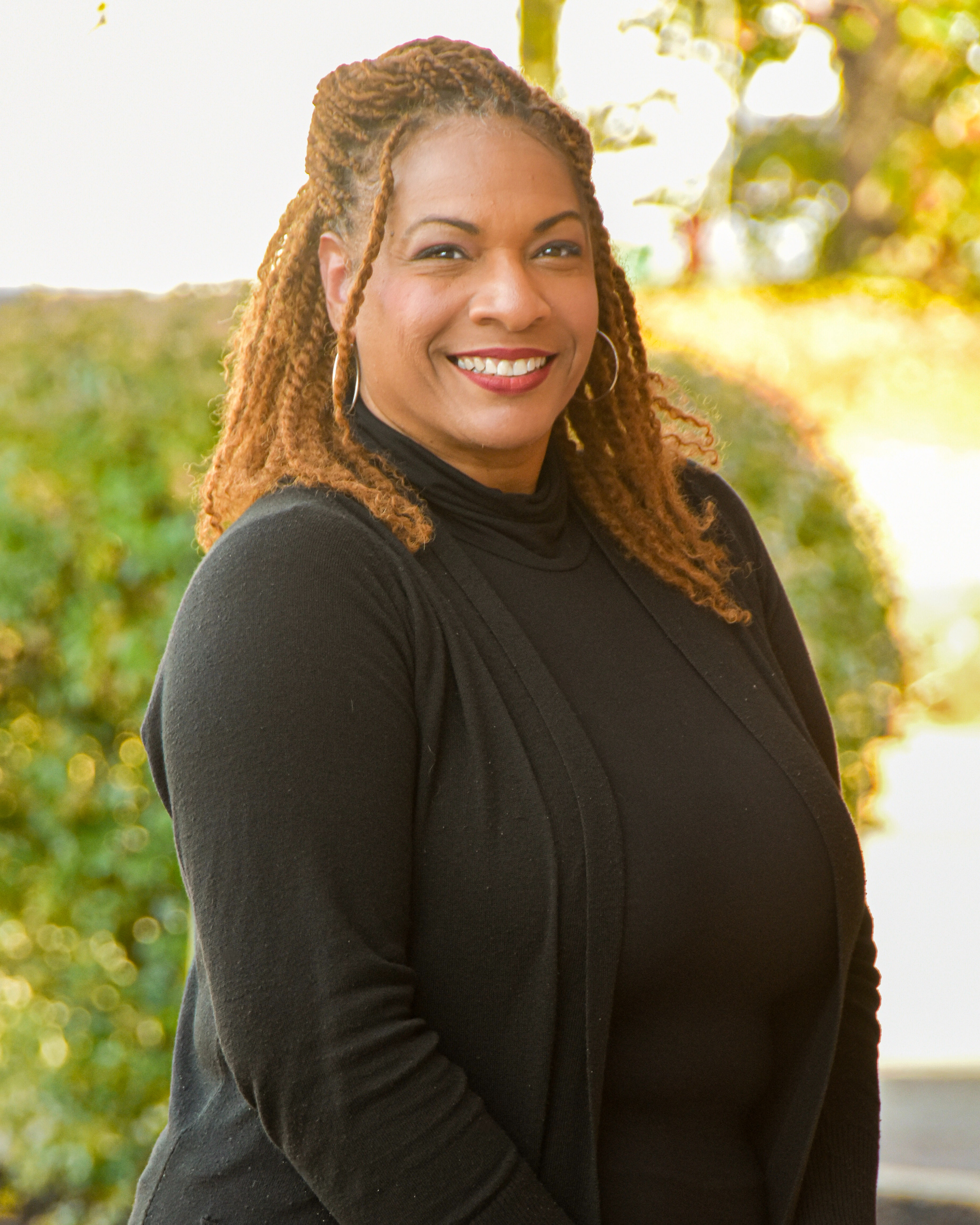A person with curly hair smiles at the camera. They wear a black turtleneck top. Behind them, greenery and a concrete sidewalk are visible.