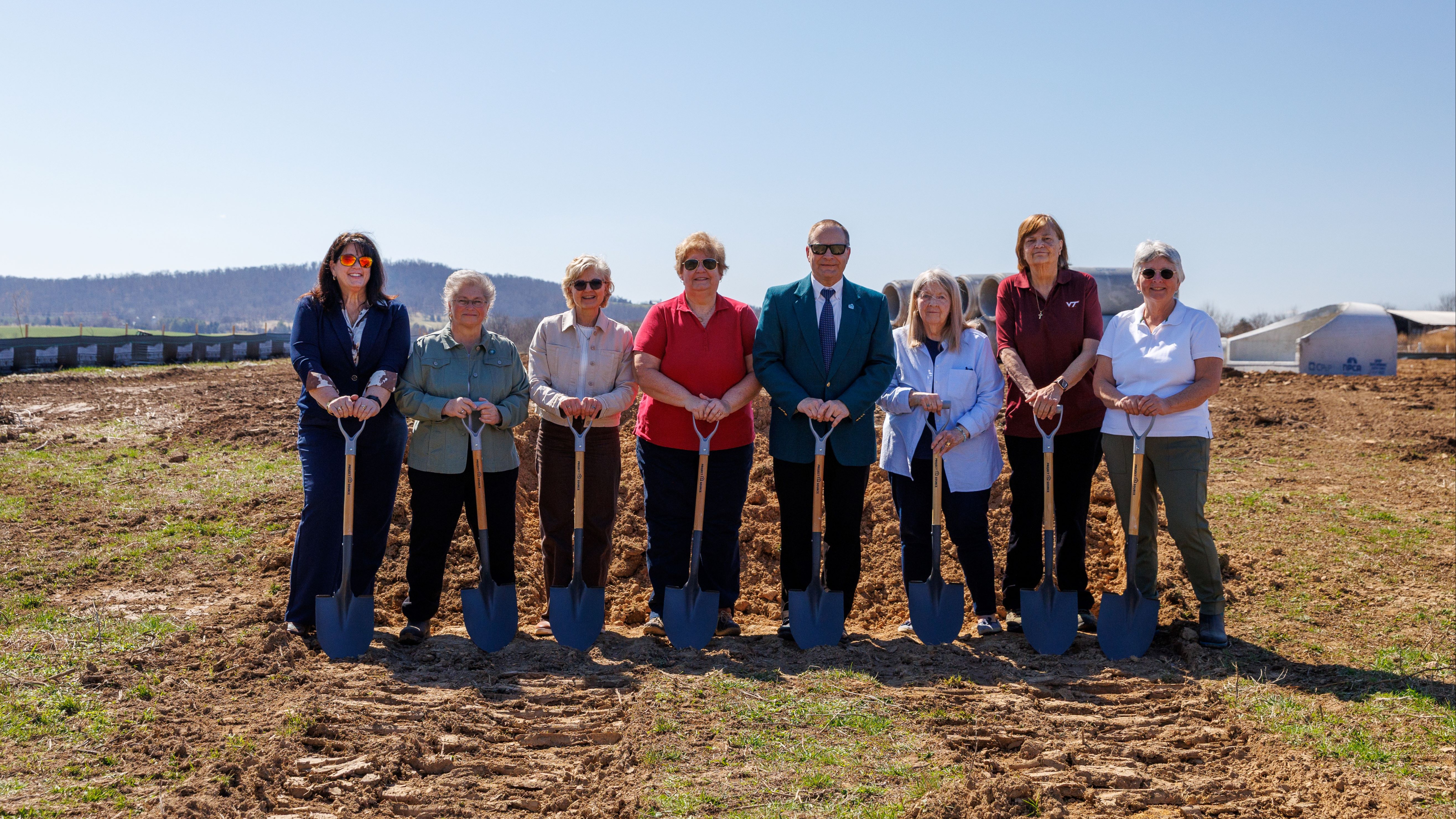 Eight people stand in a field holding shovels, ready for a ground-breaking ceremony. A blue sky with distant mountains.