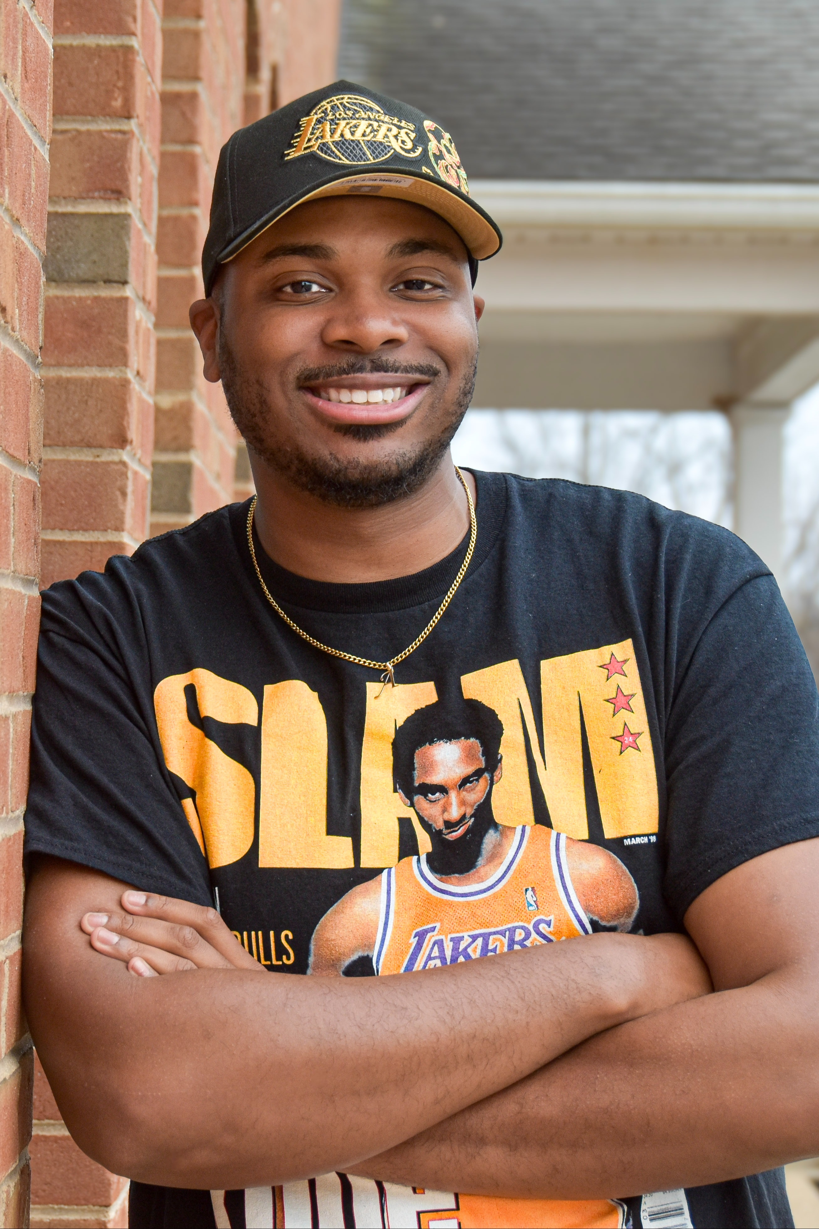 Man leaning on a brick wall with a basketball player on his shirt, smiling. Behind him, a house with a gray roof and white porch.