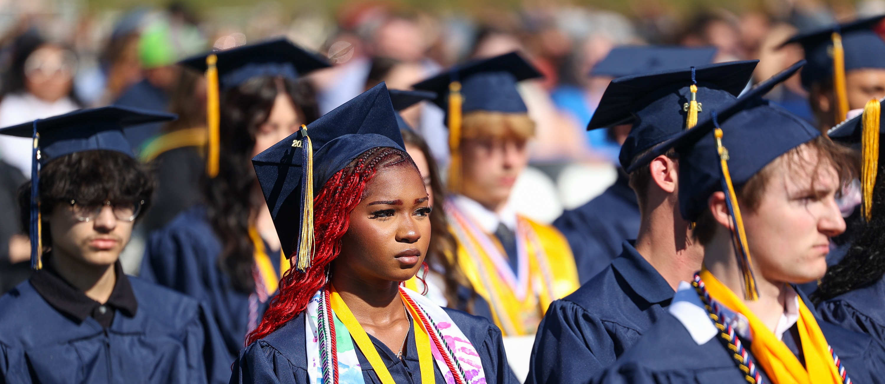 A group of young people, wearing graduation gowns and caps, attend a ceremony. They sit in rows.