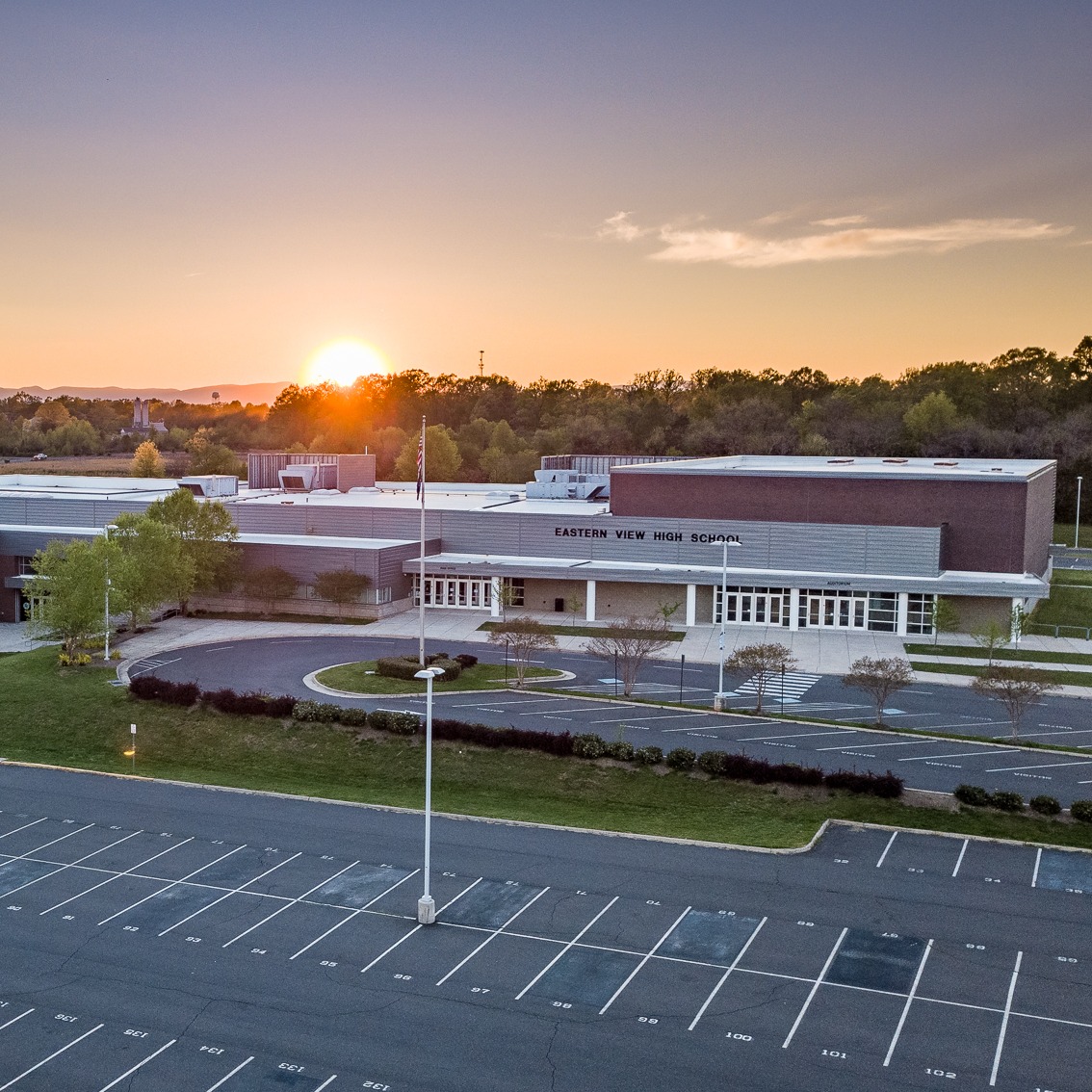A school building with a sun setting in the background, surrounded by a large parking lot.