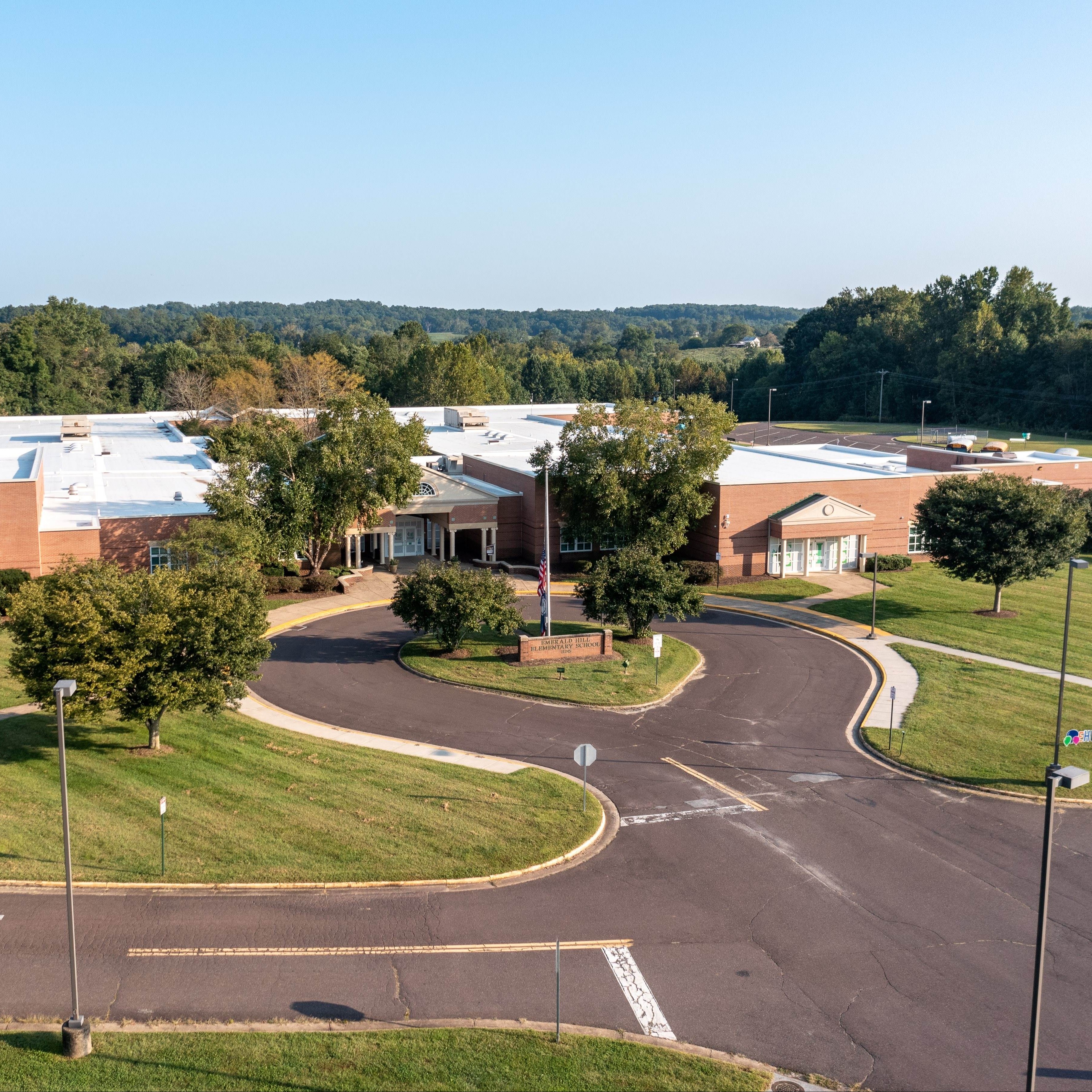 Aerial view of a building with a large parking lot, green grass, and trees. A road curves into the lot.