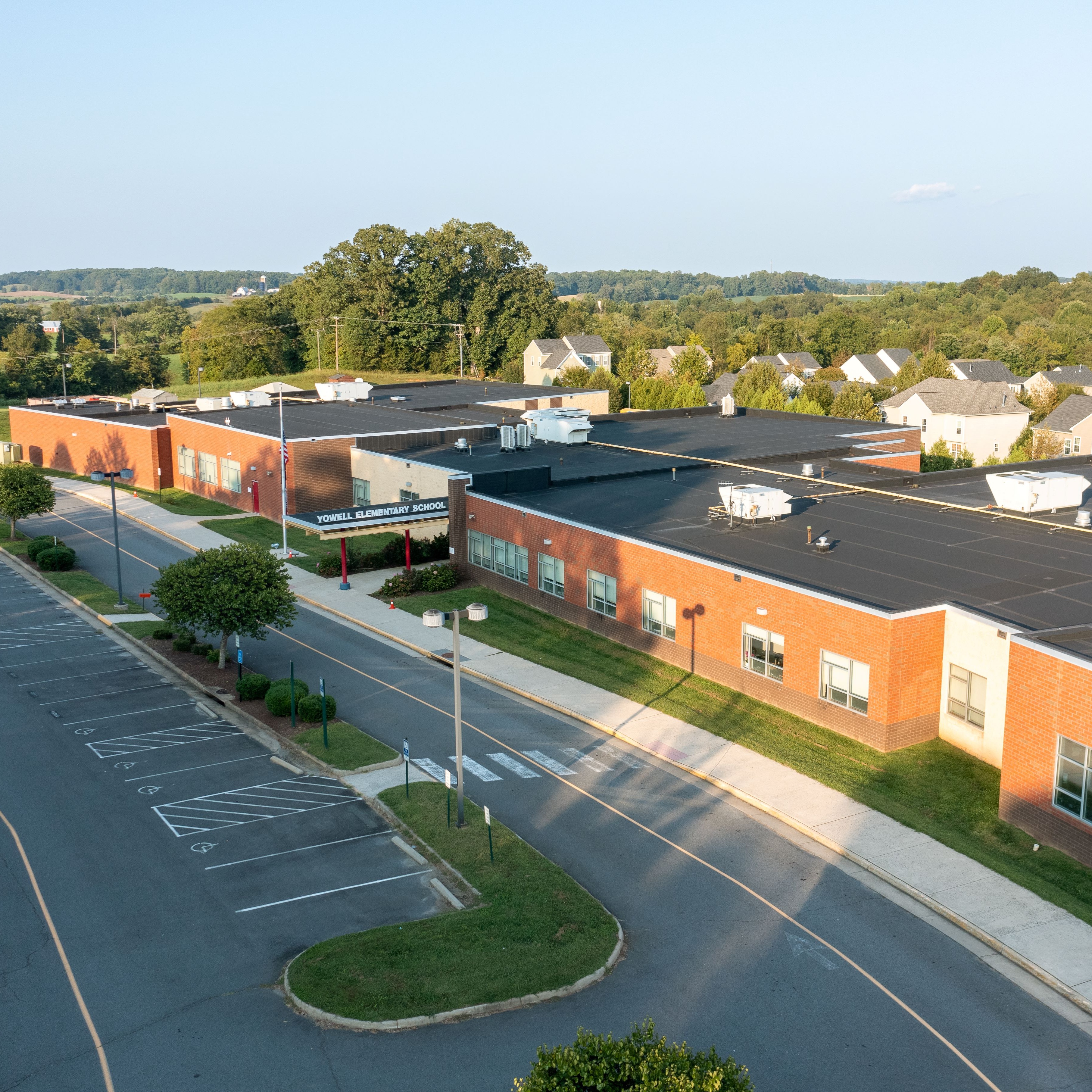 Aerial view of a large building with a parking lot, surrounded by houses, trees, and a grassy field.