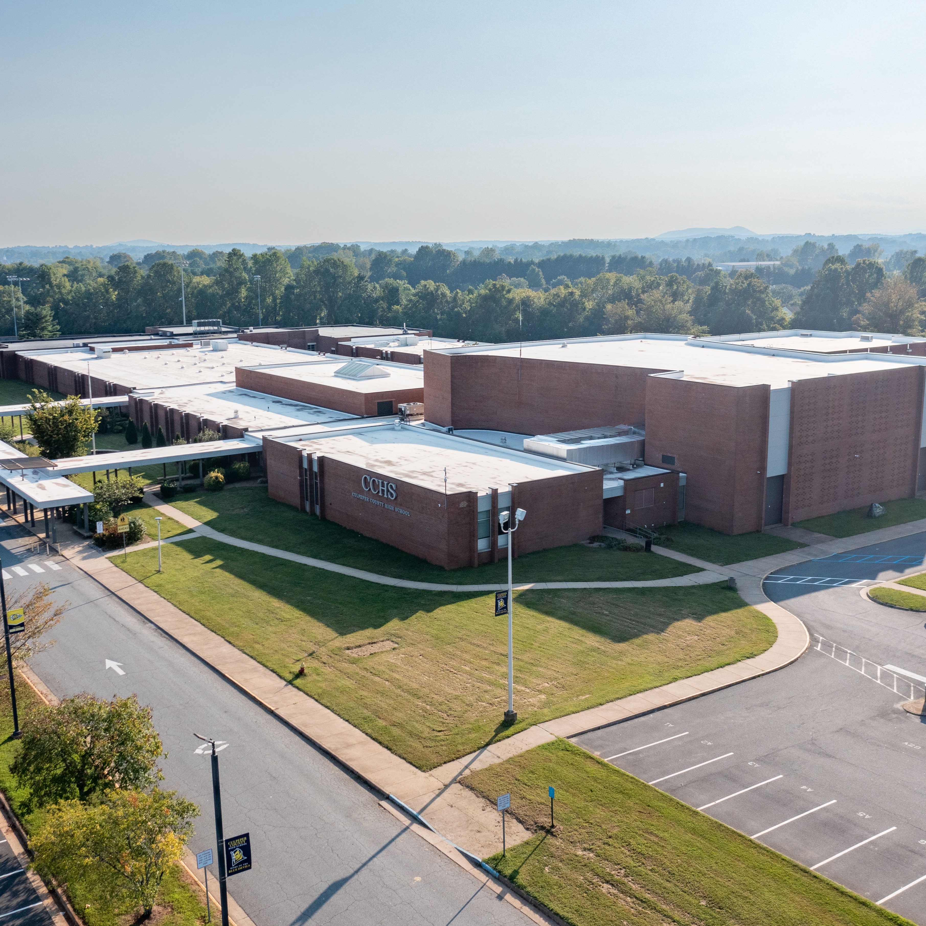 A school building with a parking lot and a road in front of it. Green grass and trees surround the building.