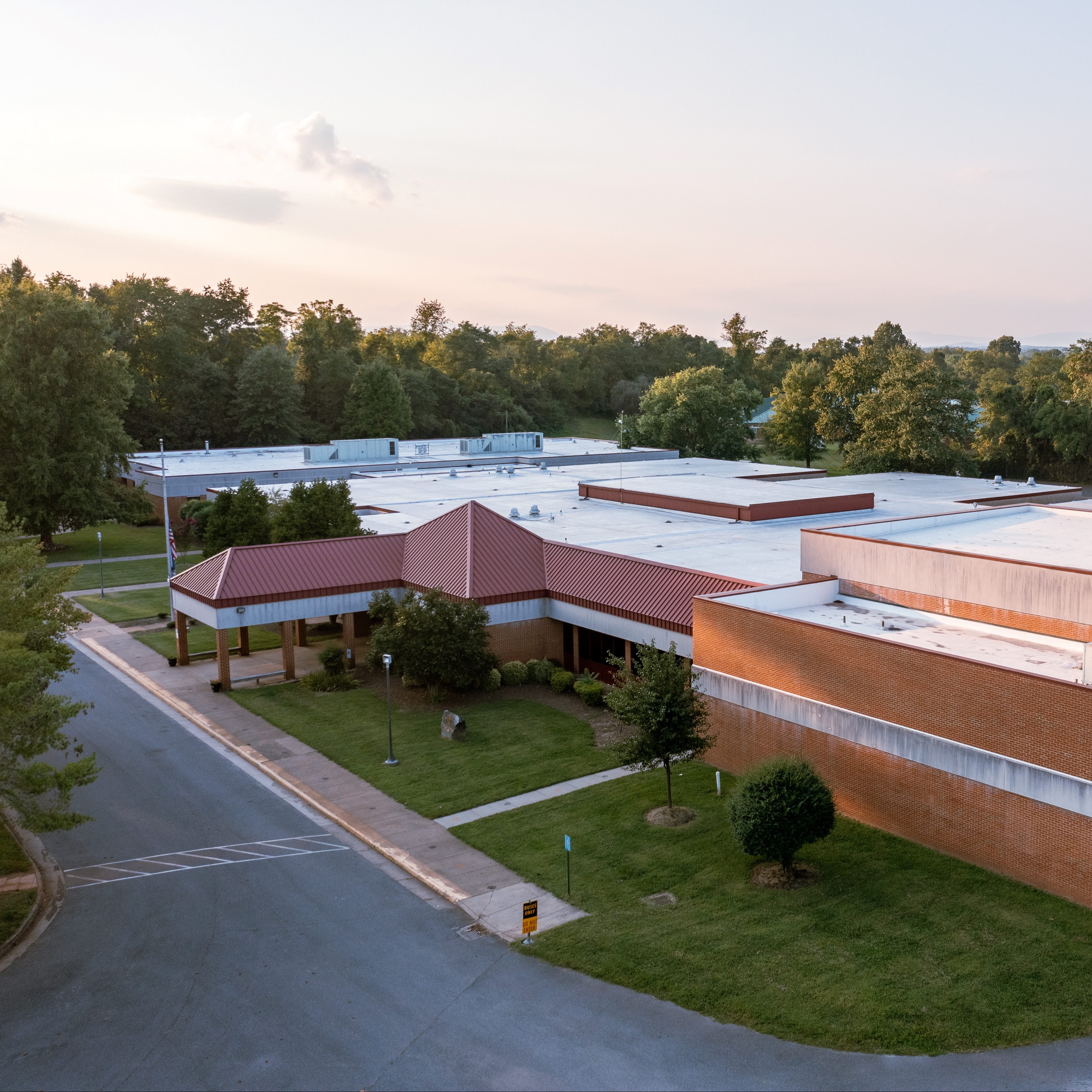 Aerial view of a building with a red roof and a white roof, surrounded by trees and grass.