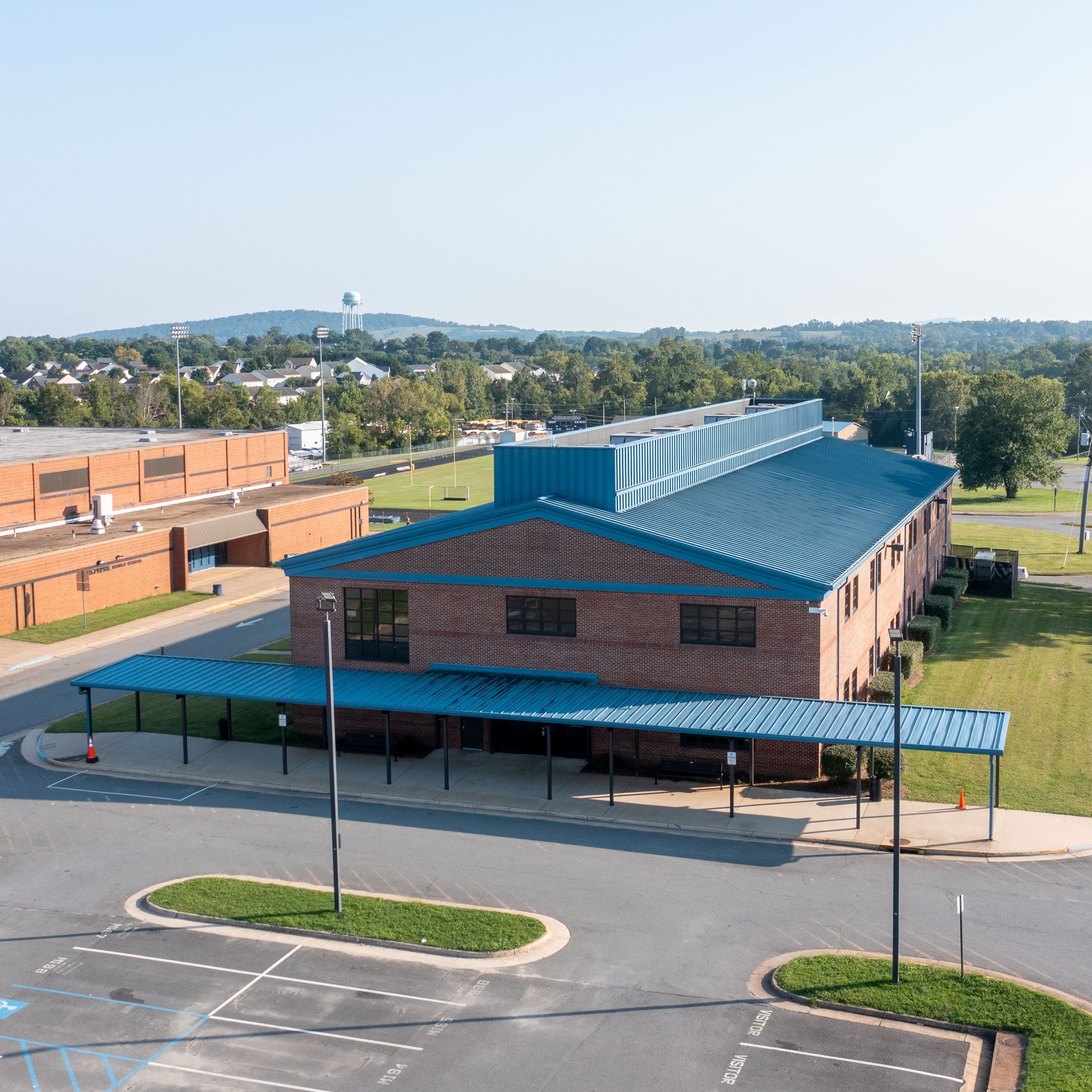 A school building with a blue roof and a parking lot. Trees, grass, and a road are visible.