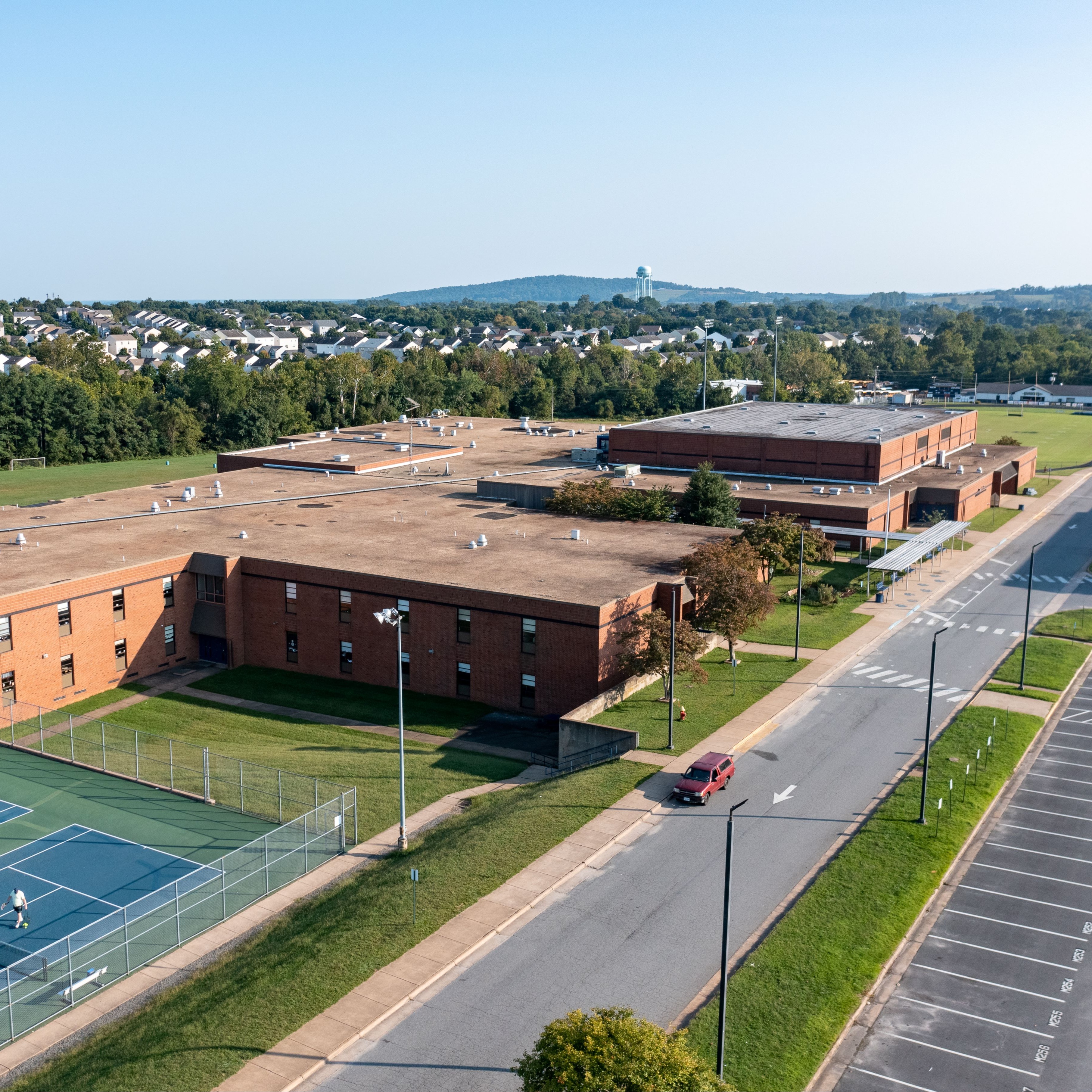Aerial view of a school building with tennis courts in front, a road, and a parking lot.