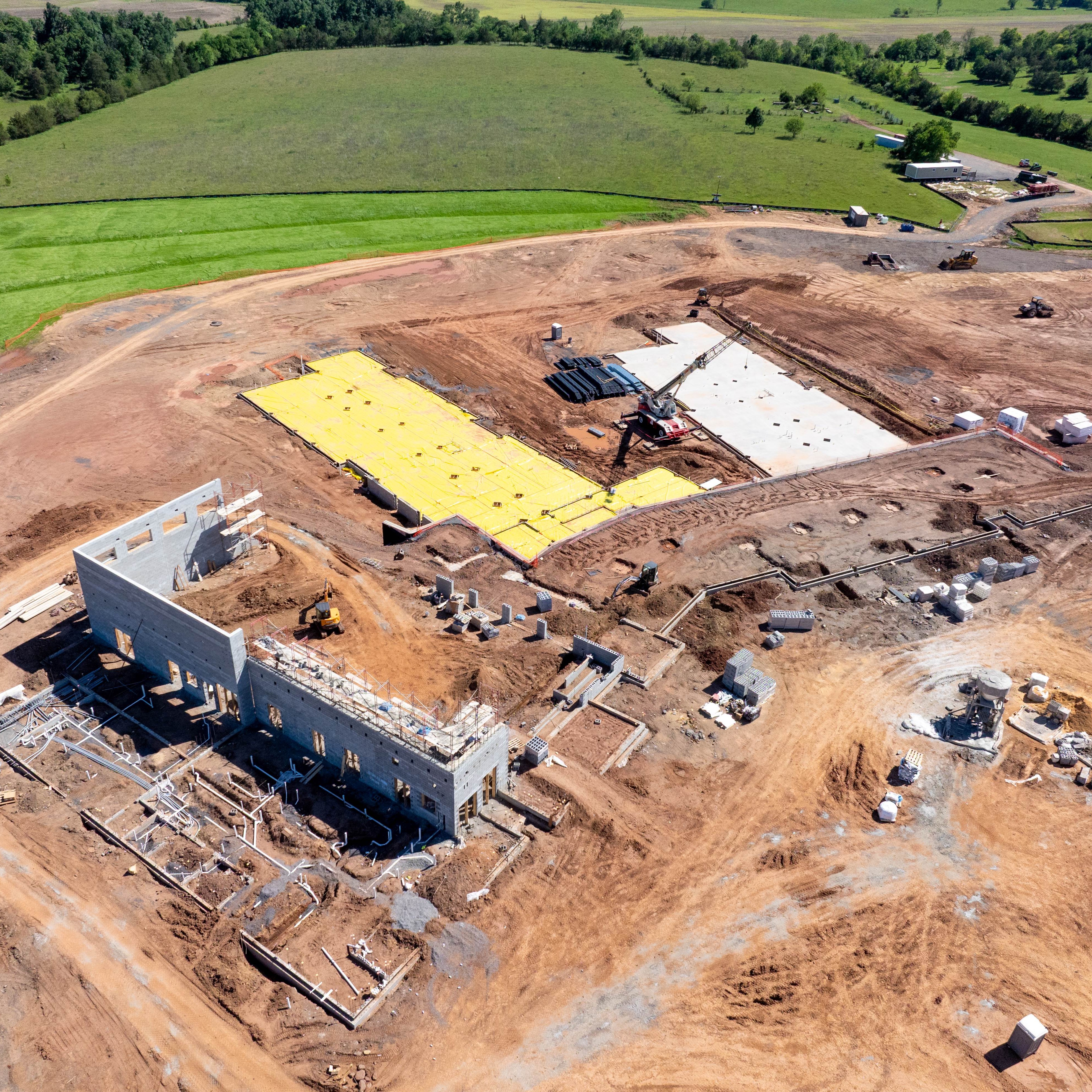 Aerial view of a construction site with a partially built building, yellow tarp, and surrounding dirt fields.