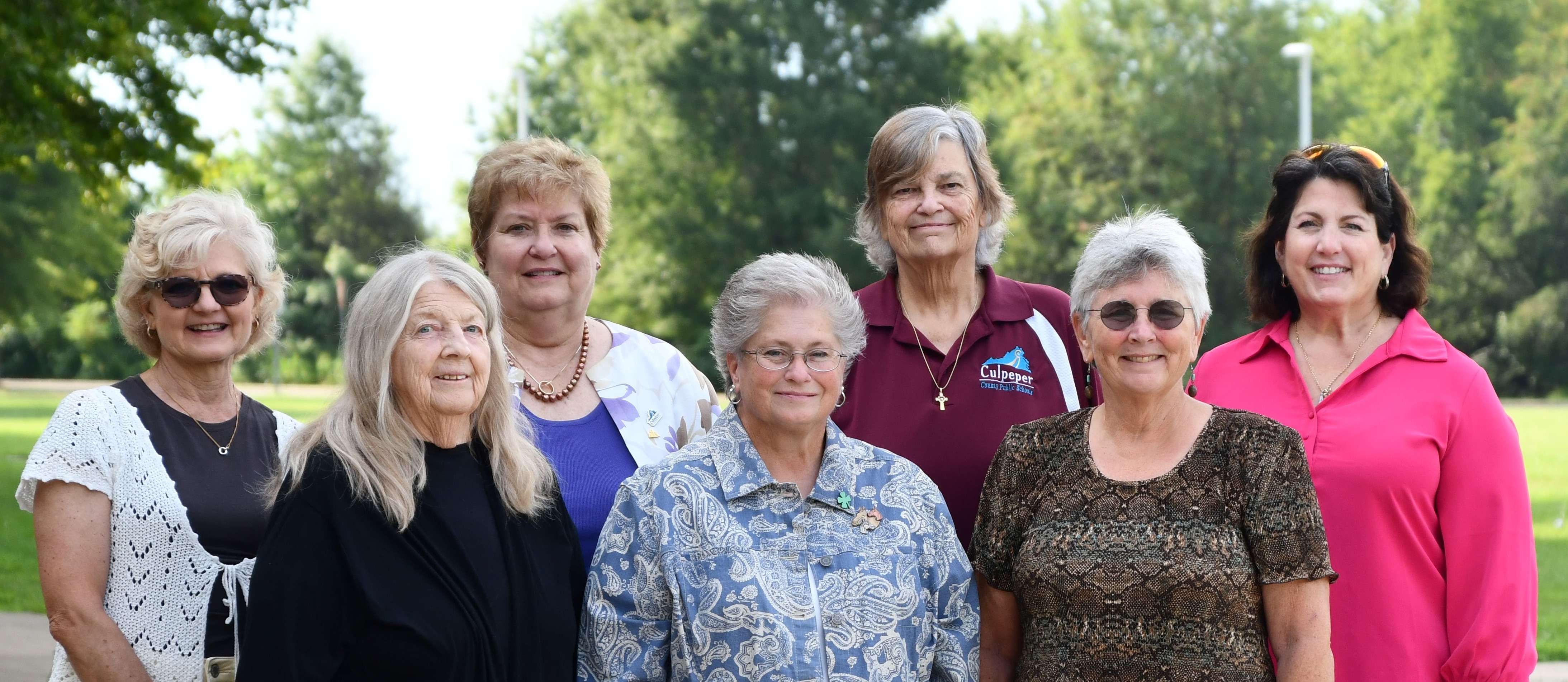 A group of women standing together on a path in a park with trees and grass.