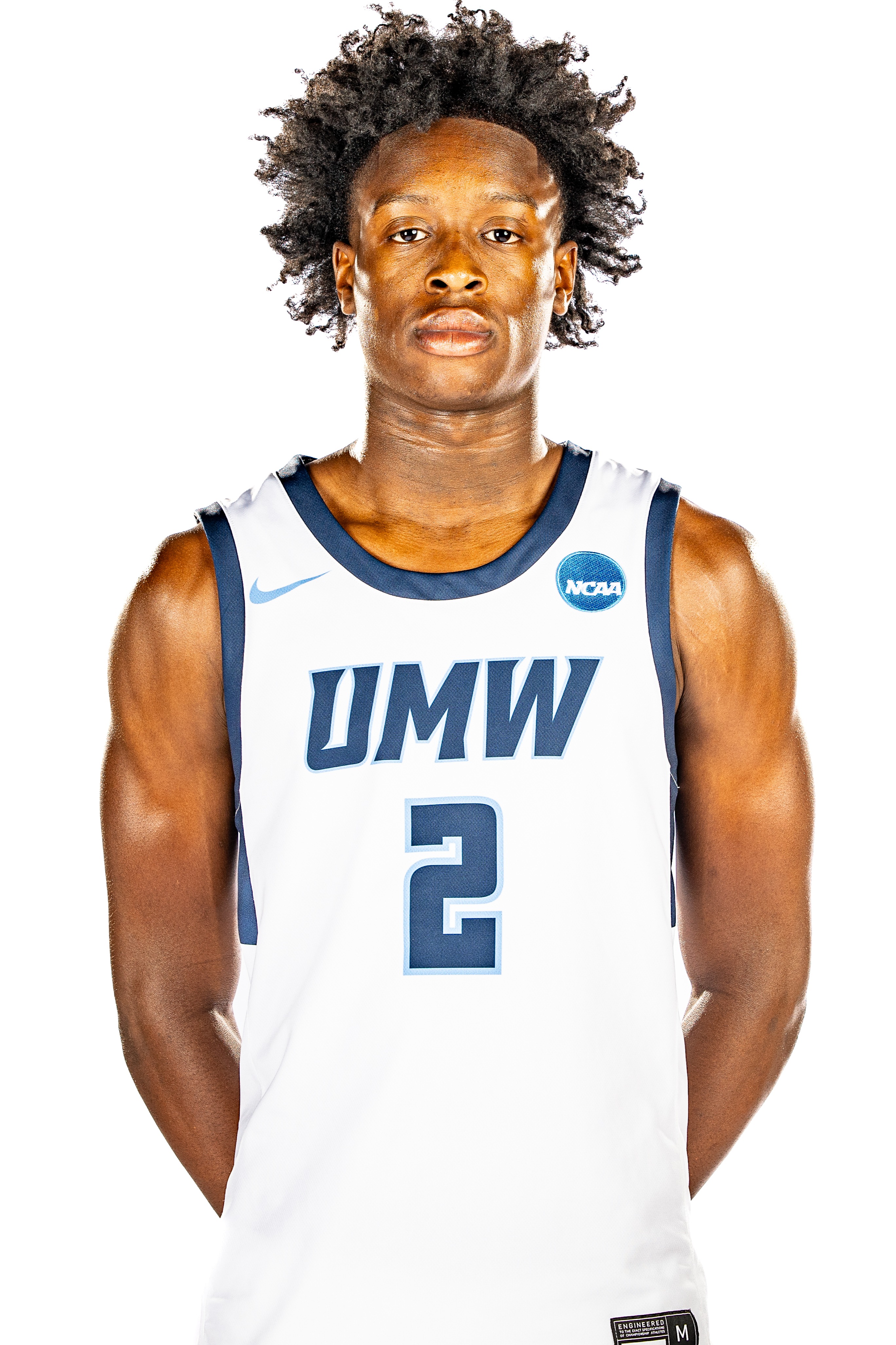 A basketball player with curly hair stands with hands on hips. He wears a white and blue uniform with UMW and number 2.