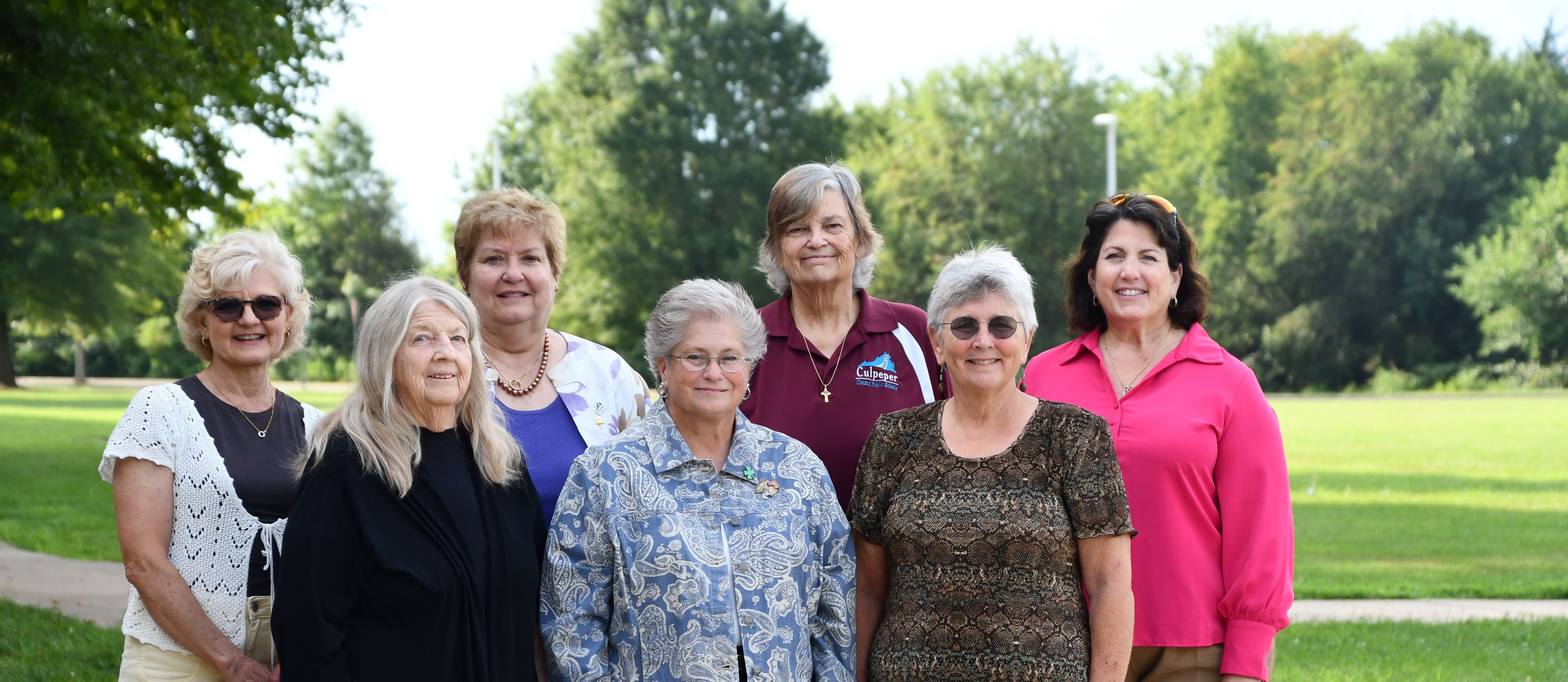 A group of women, dressed in various colors, standing together in front of a green park.