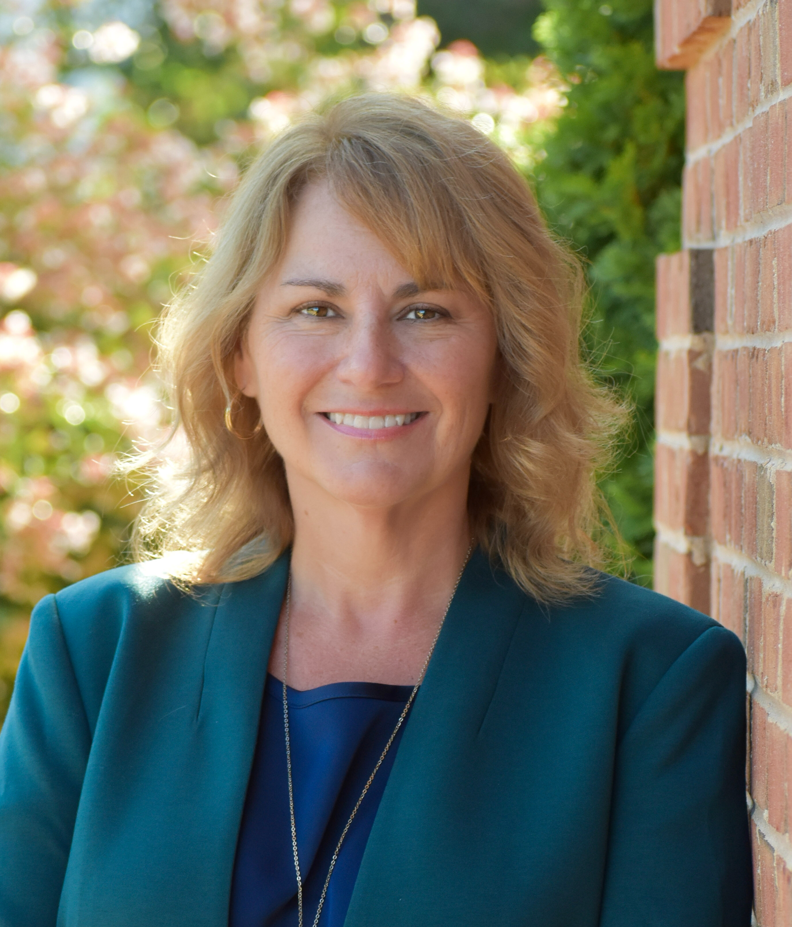 Woman with blonde hair in a teal blazer. She smiles at the camera. Blurry green plants behind her.