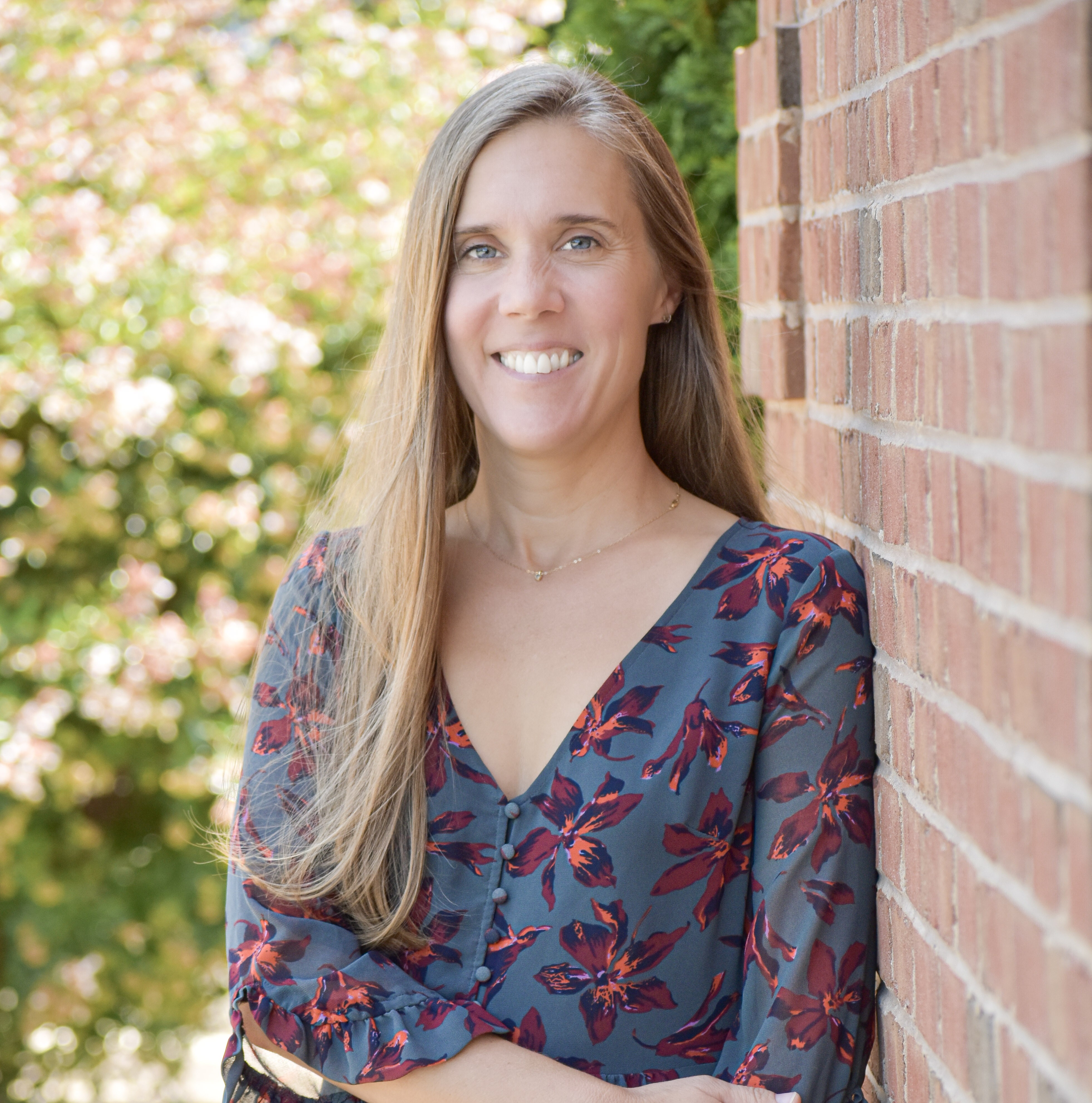Woman with long blonde hair leaning against a brick wall, smiling, and wearing a floral blouse.