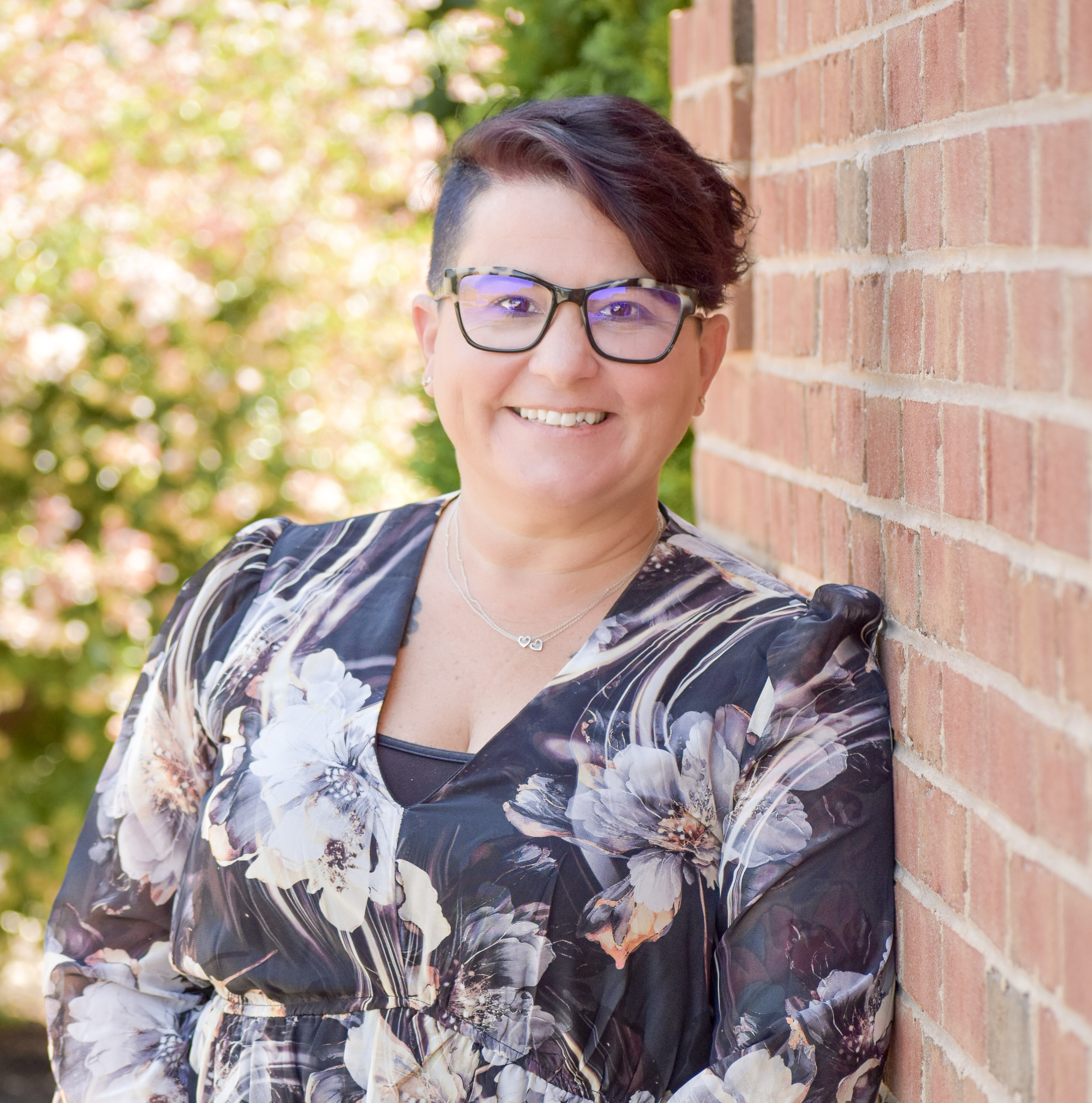 A woman wearing glasses, a floral dress, and a necklace smiles, leaning against a brick wall with pink flowers.