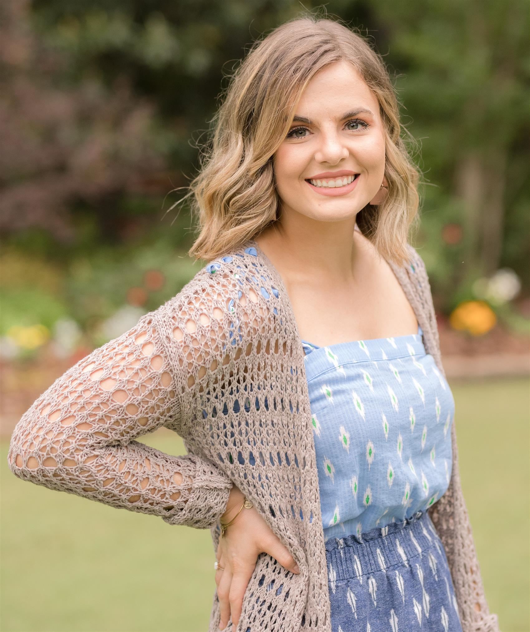 Woman wearing blue top and light colored cardigan sweater in front of green tree setting