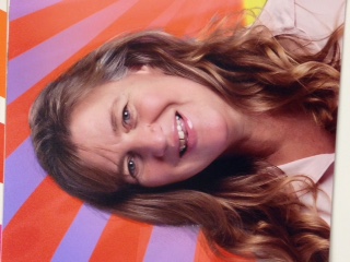 Smiling woman with brown hair in front of purple and orange sunbeam background.