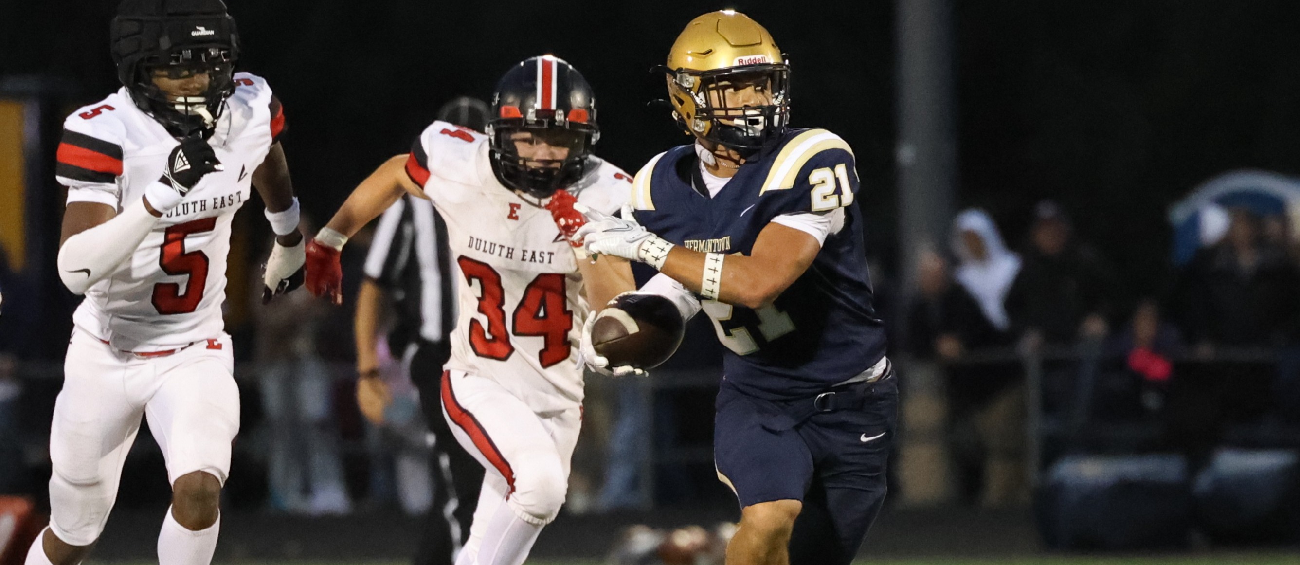Three football players in helmets and sneakers run on a field. The player in blue wears a gold helmet.