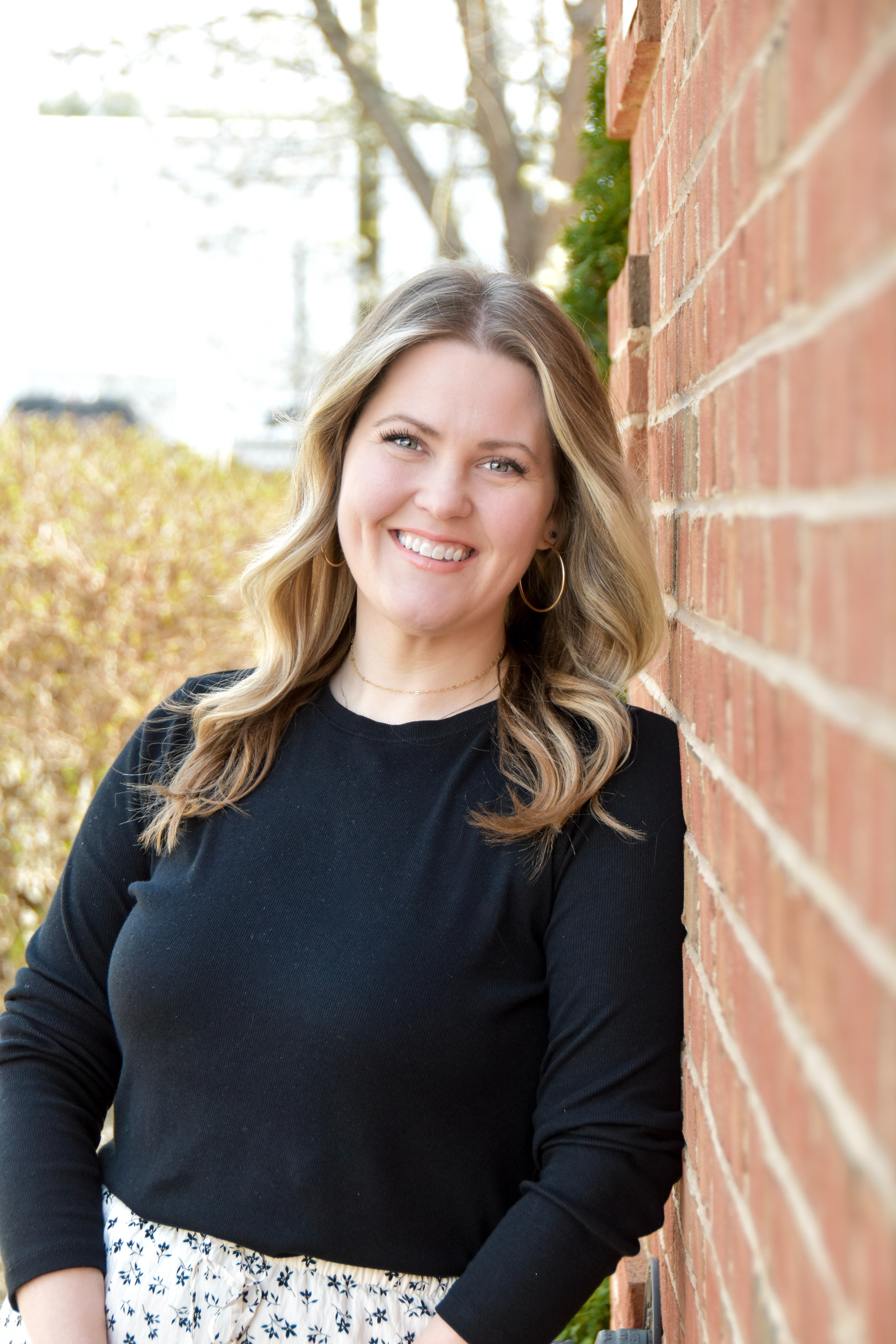 A smiling woman with blonde hair leans against a brick wall, wearing a black long-sleeve shirt and a floral skirt.