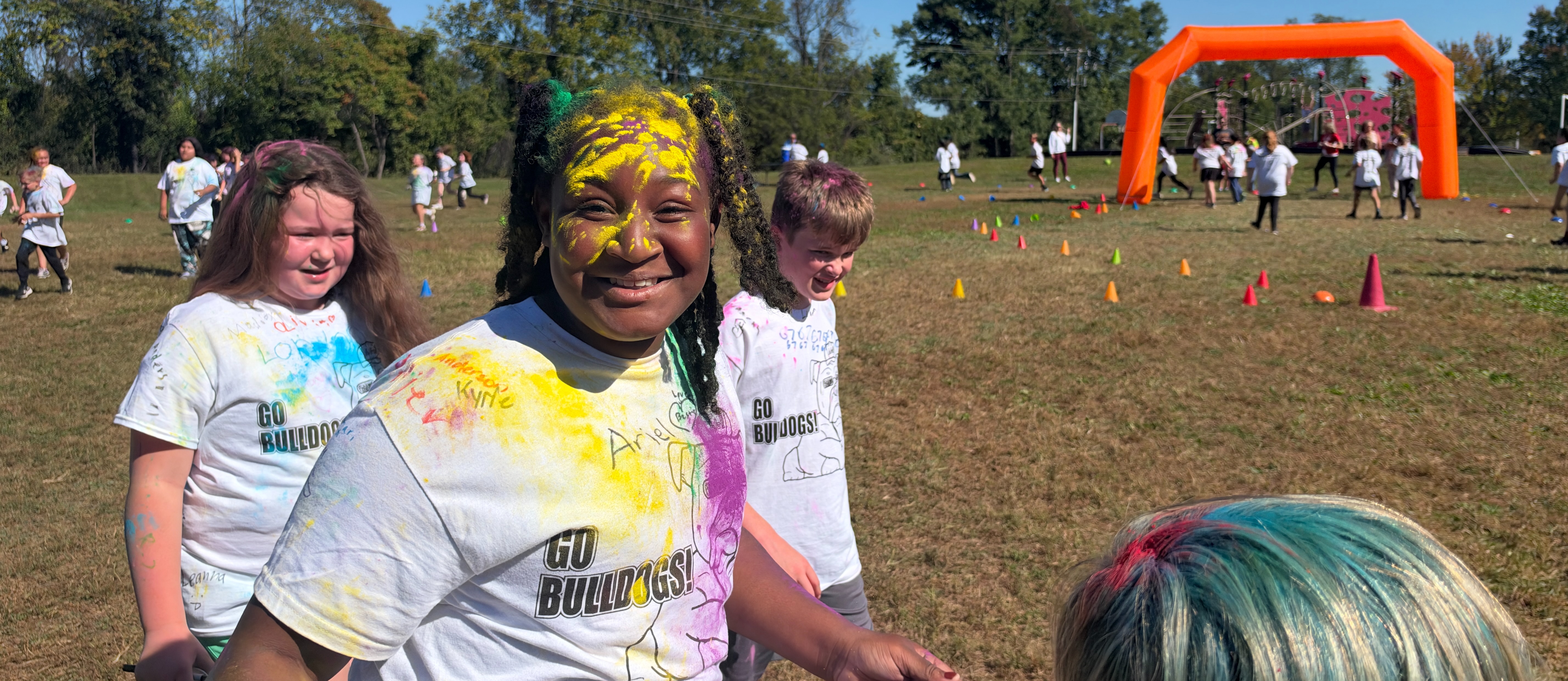 People wearing colorful shirts stand in a field, smiling. One person has yellow and pink on their face. Orange arch and traffic cones are in the background.