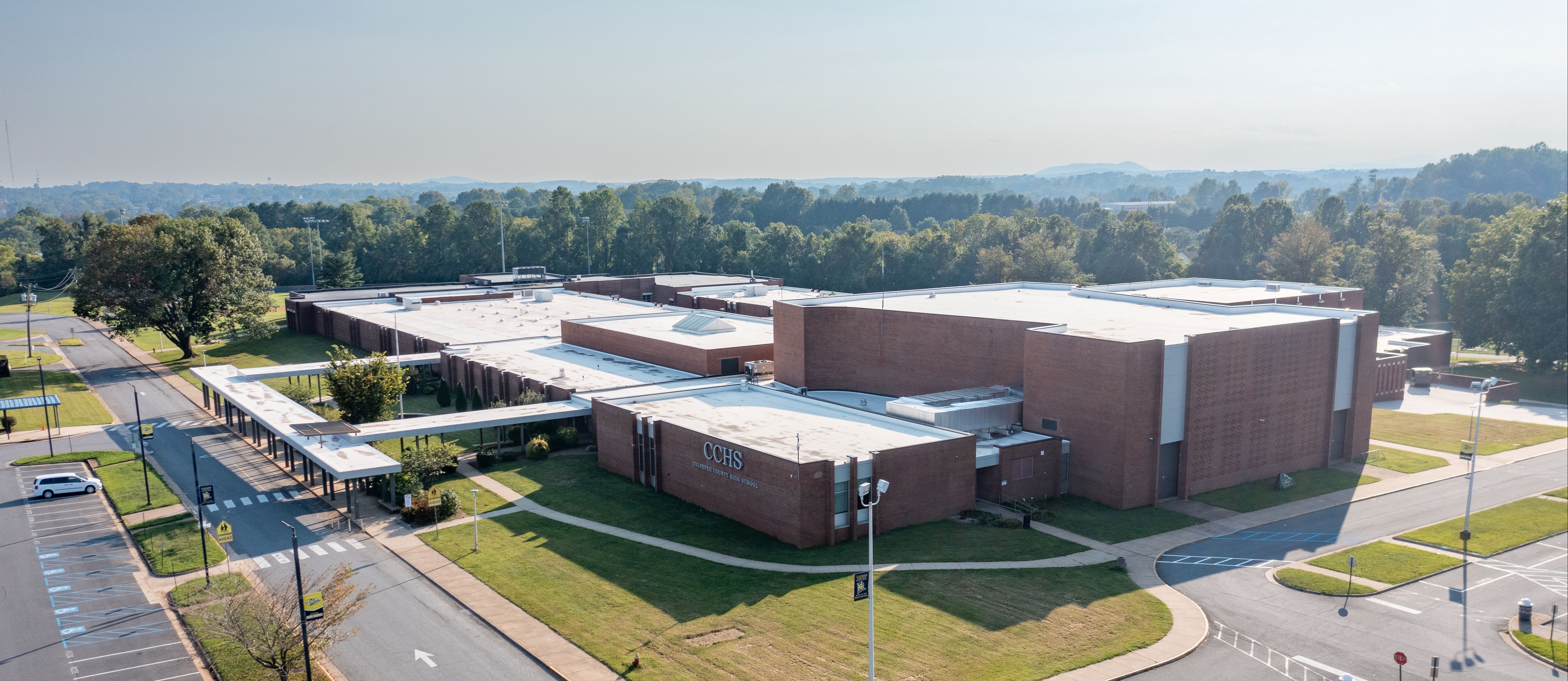 Aerial view of a large, brown building with a flat roof. Green lawns and parking lot with cars parked. Trees and mountains in the distance.
