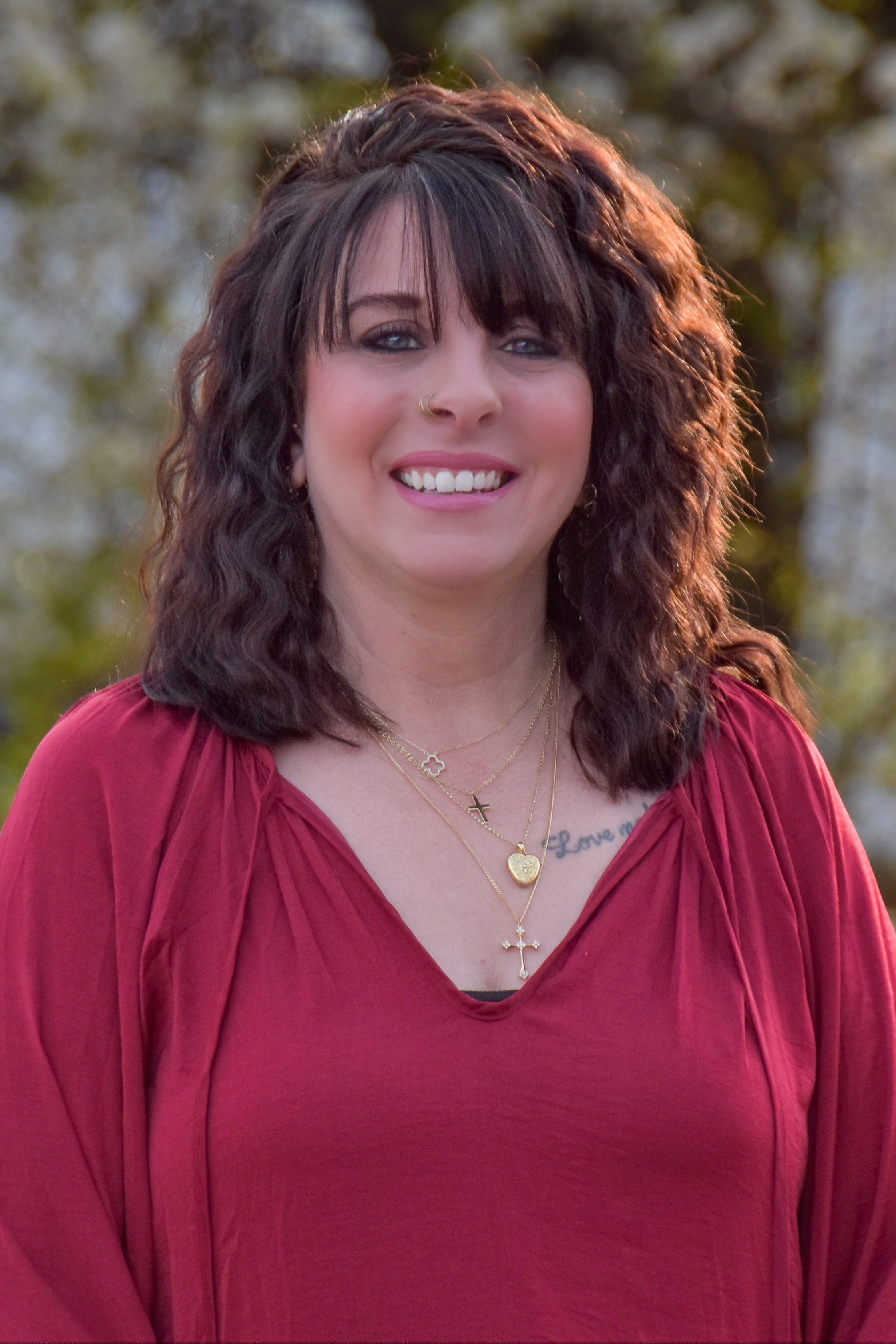 A smiling woman with brown curly hair stands in a park. She wears a red blouse and necklace.