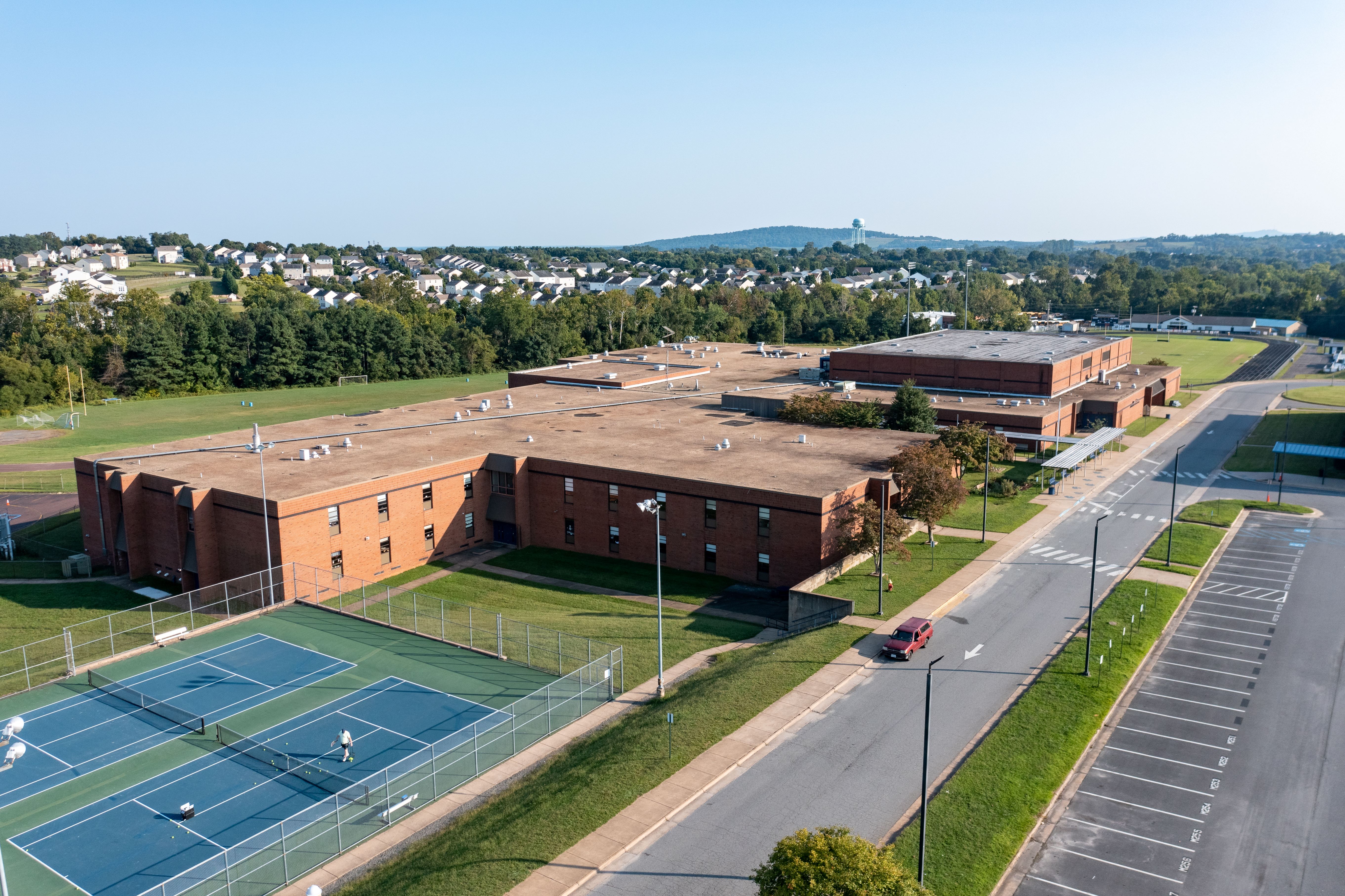 Overhead view of a school building, tennis courts, parking lot, trees, and hills in the distance.