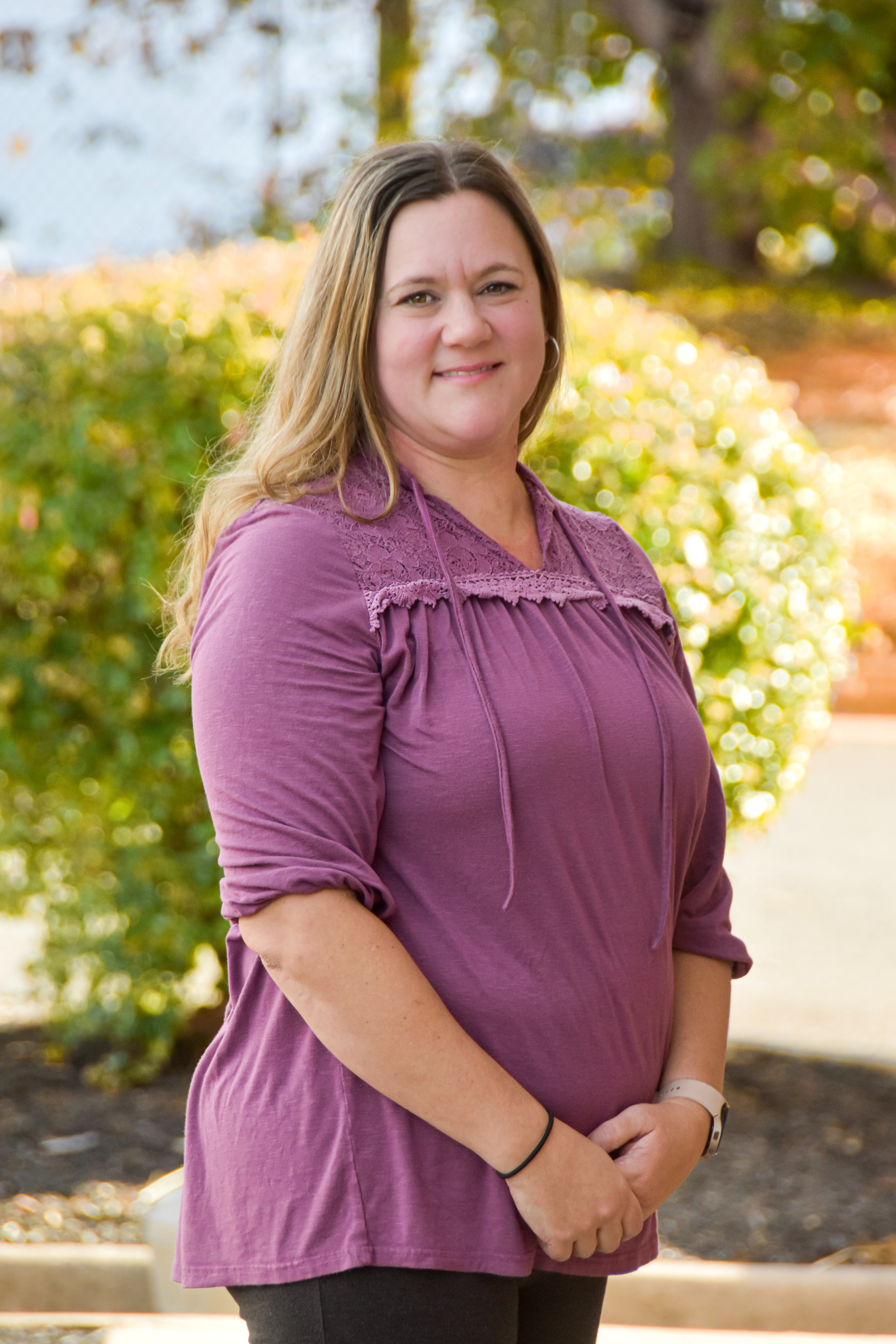 A woman stands outside, smiling, in a purple blouse and black pants. Green shrubs and trees are in the background.