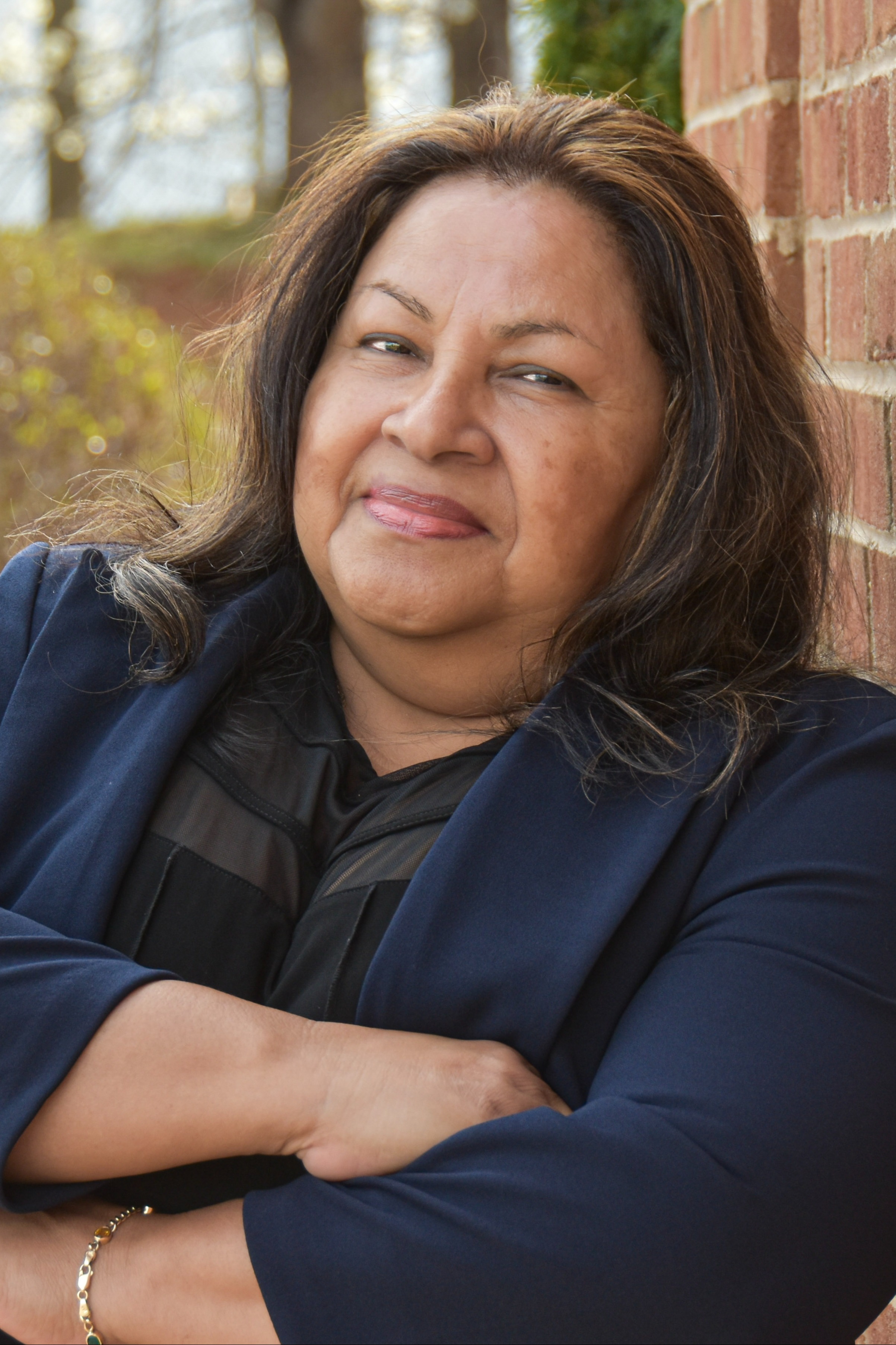 An adult woman stands against a brick wall with her arms crossed. She wears a blue jacket and black top. Behind her is greenery.