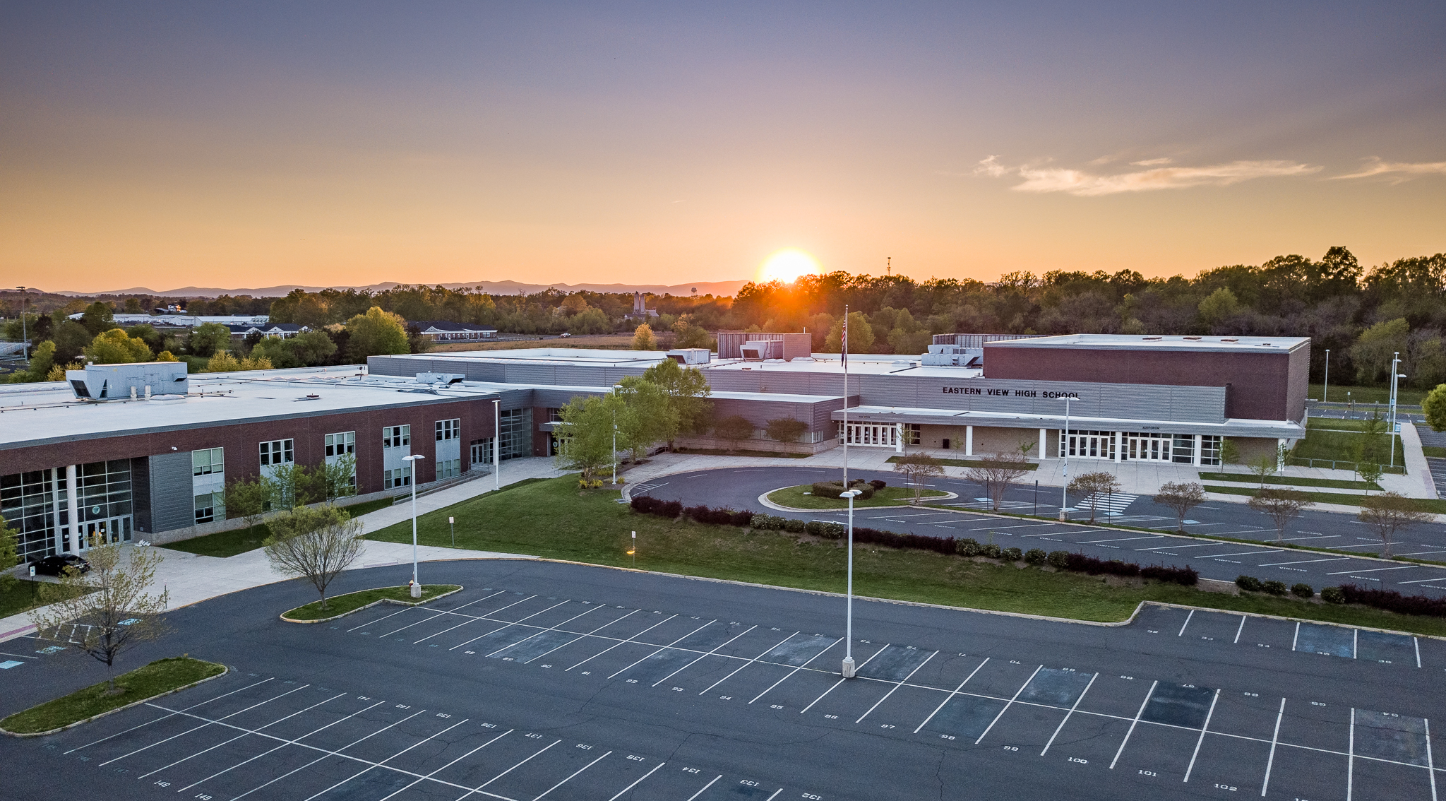 An aerial view of a modern school building with a parking lot and trees. The sun is setting.