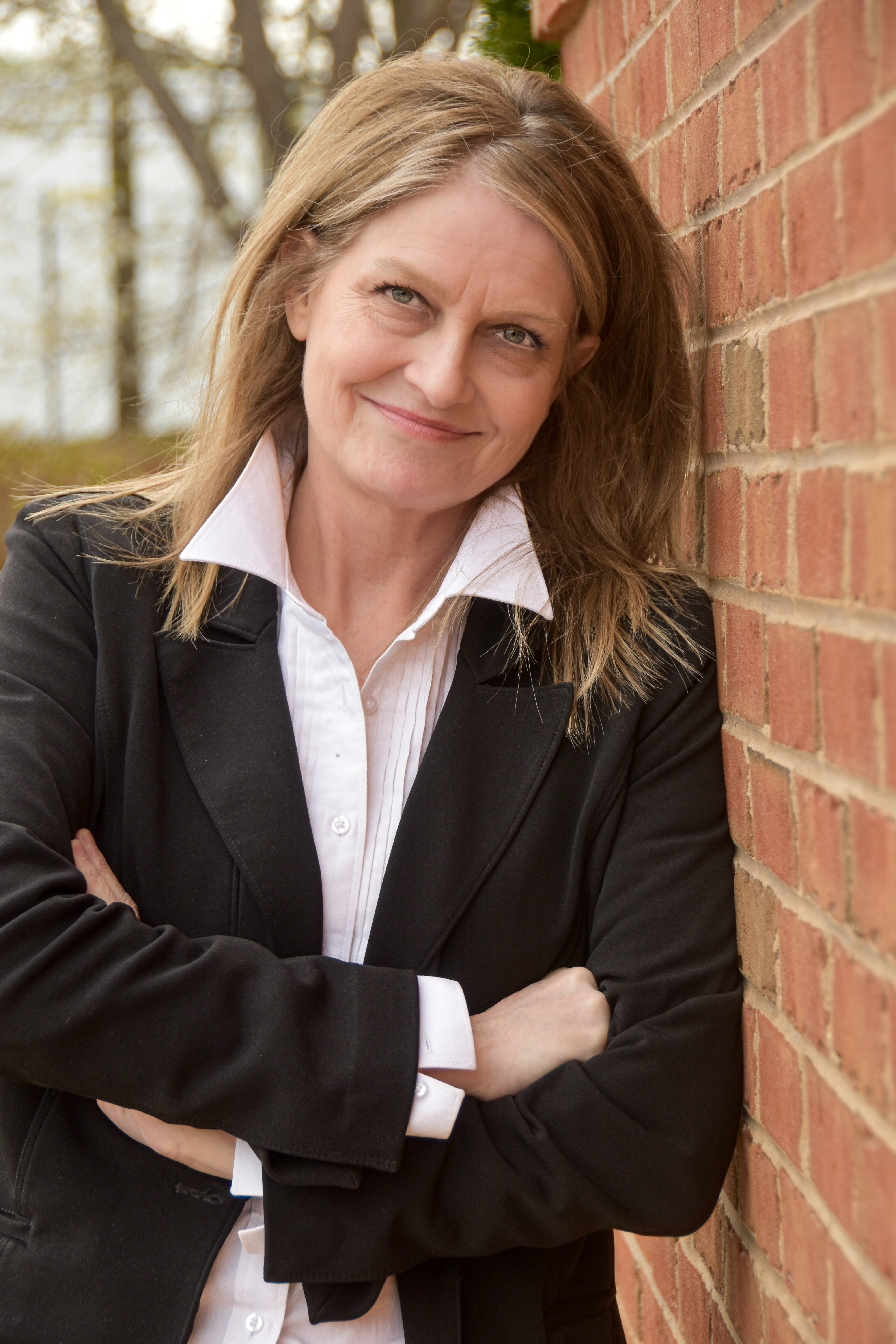 A woman with blonde hair wearing a black blazer and white shirt leaning against a brick wall, smiling.