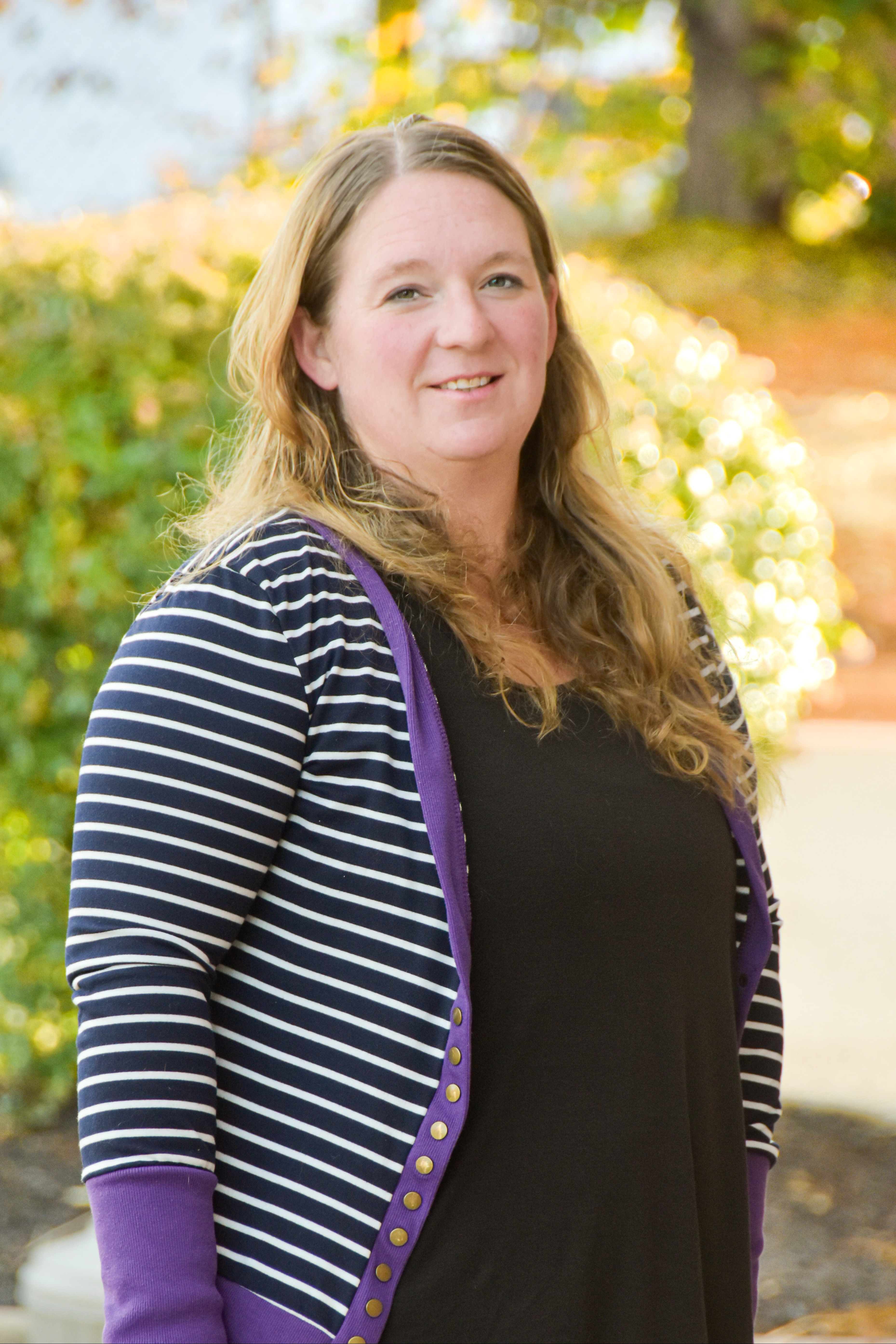 A woman with blonde hair, wearing a striped cardigan and a black top, stands smiling in front of greenery.