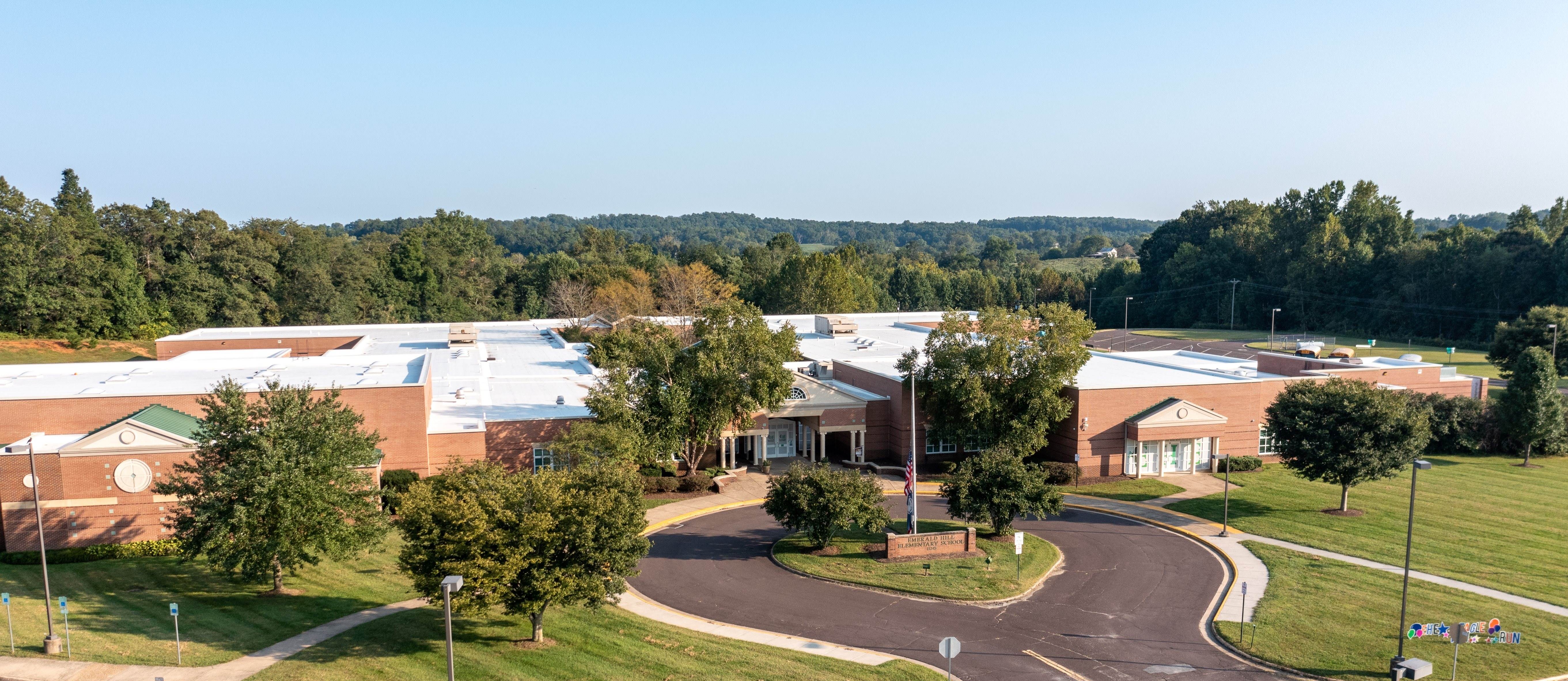 Aerial view of a school building with a parking lot, trees, and a grassy field. The building is surrounded by greenery.