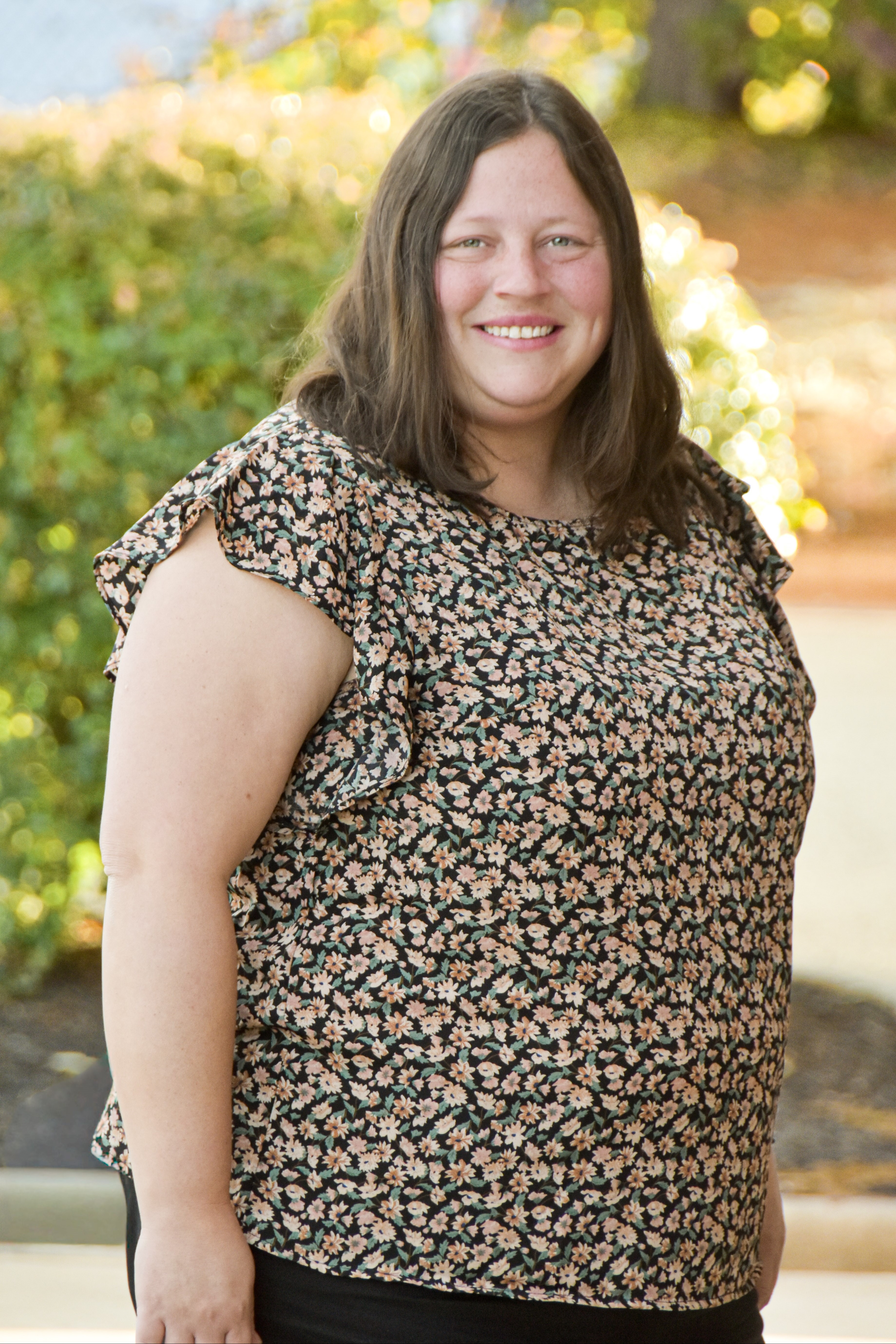 A woman with brown hair smiles in front of green plants, wearing a floral blouse with ruffles.
