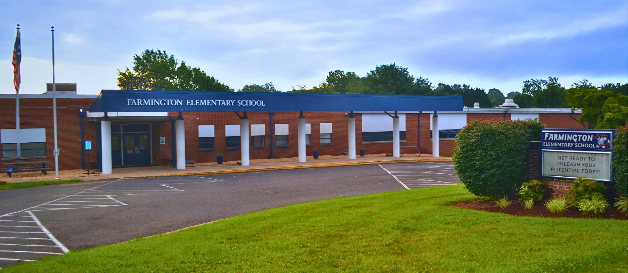 Front of a brick building with blue roof. Green lawn, sign, bushes, and flagpole. Trees in distance.