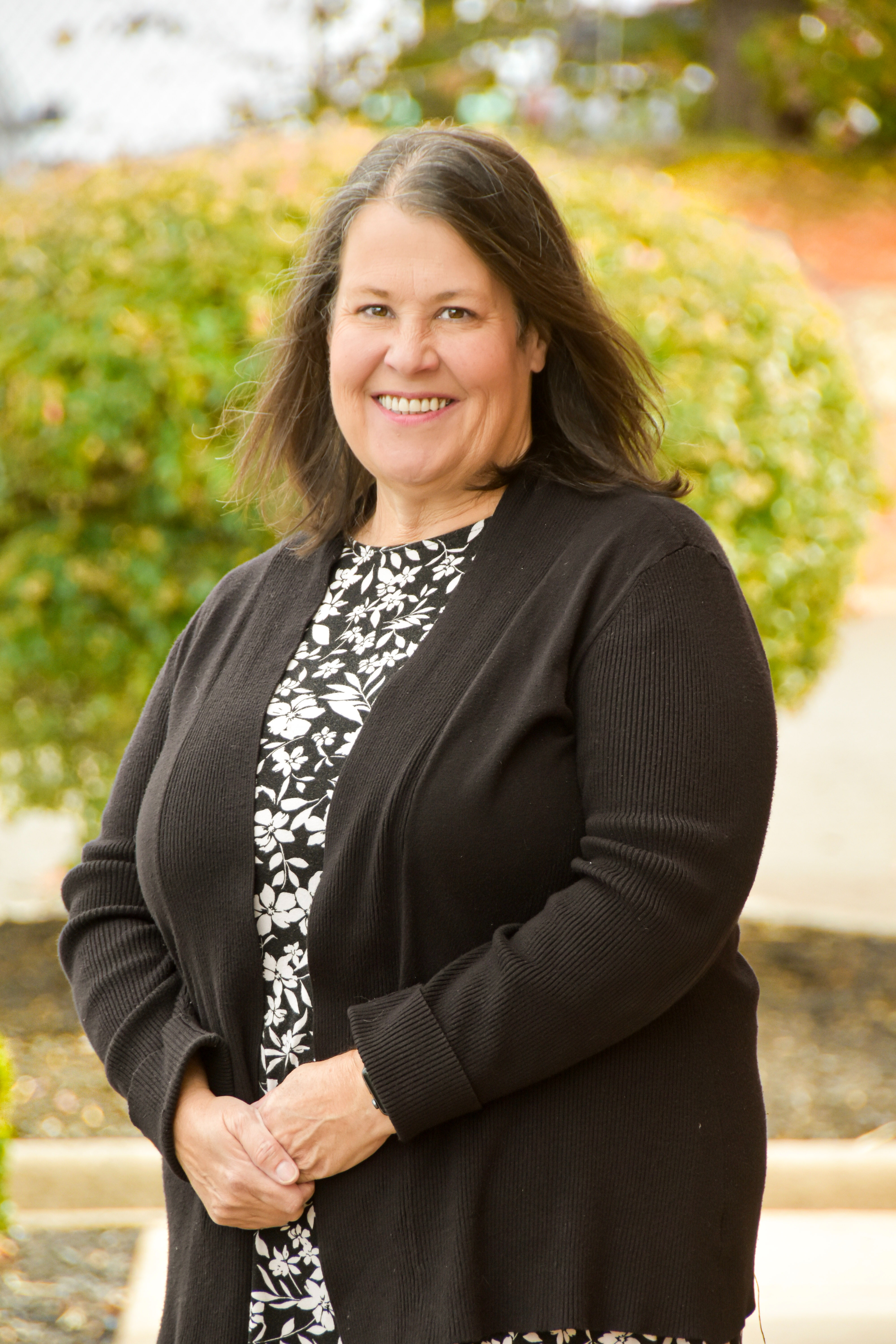Woman smiling with hands clasped, wearing a black cardigan over a patterned blouse, standing outside with green shrubs.
