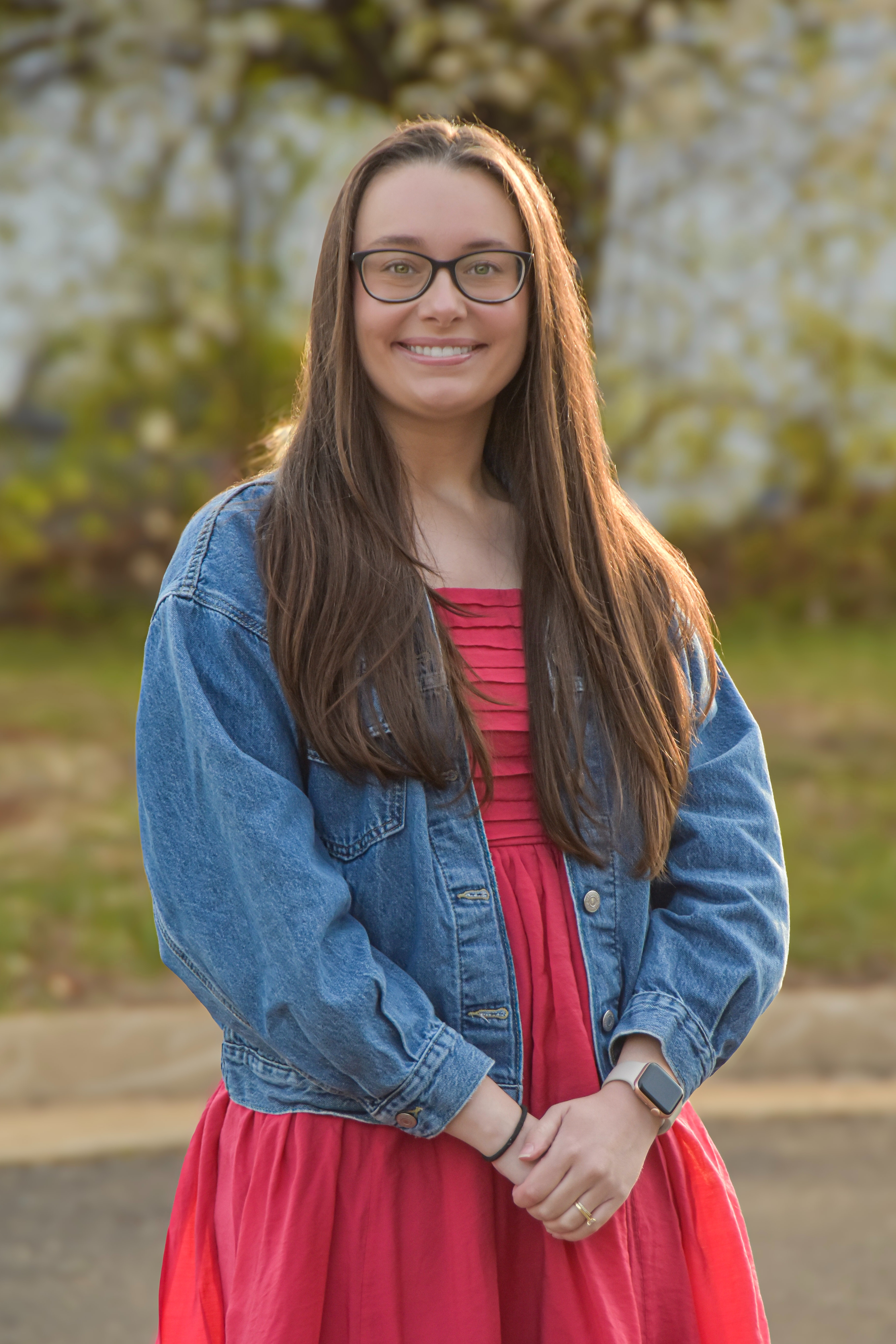 Woman in a red dress and denim jacket smiles outside. Behind her, a tree with white flowers and a white building.