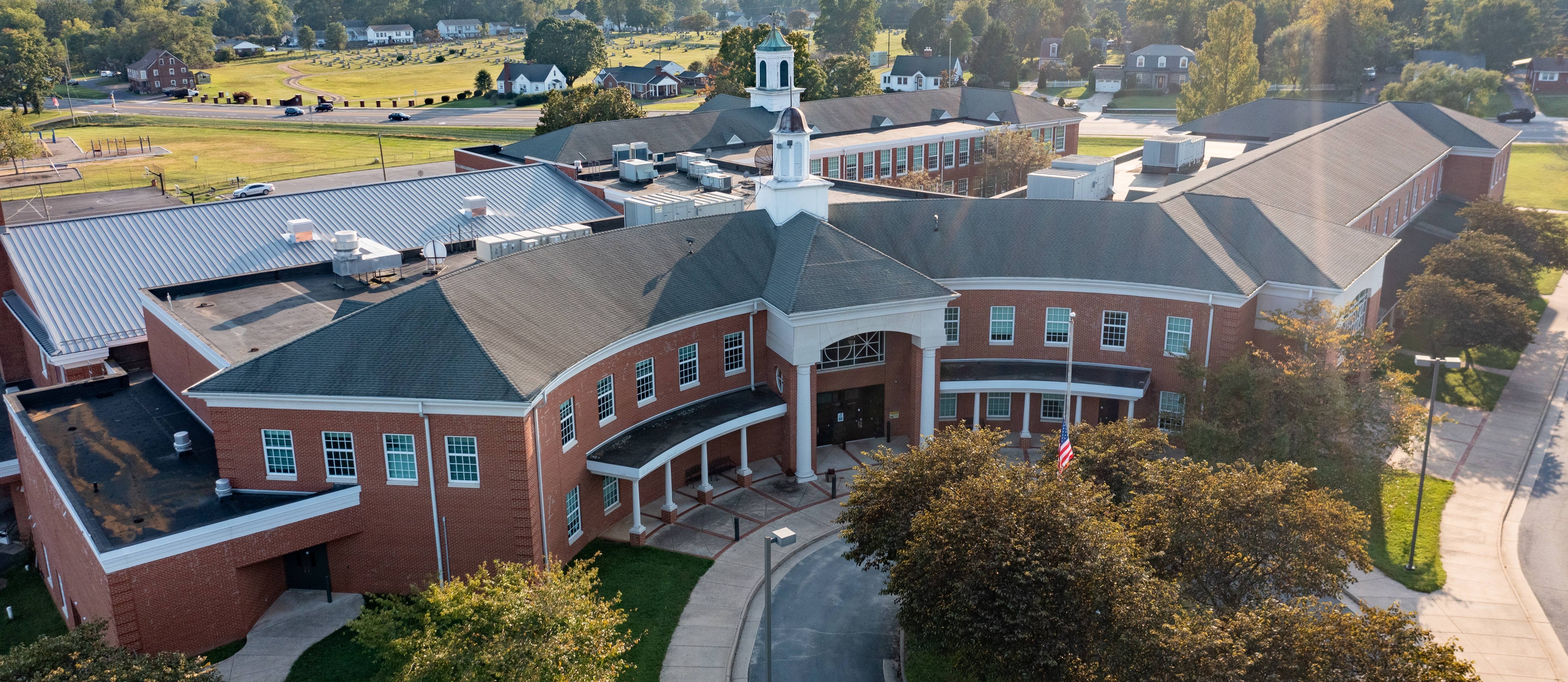 Bird's-eye view of a large brick building with a gray roof and a tower. Trees, a sidewalk, and a driveway are present.