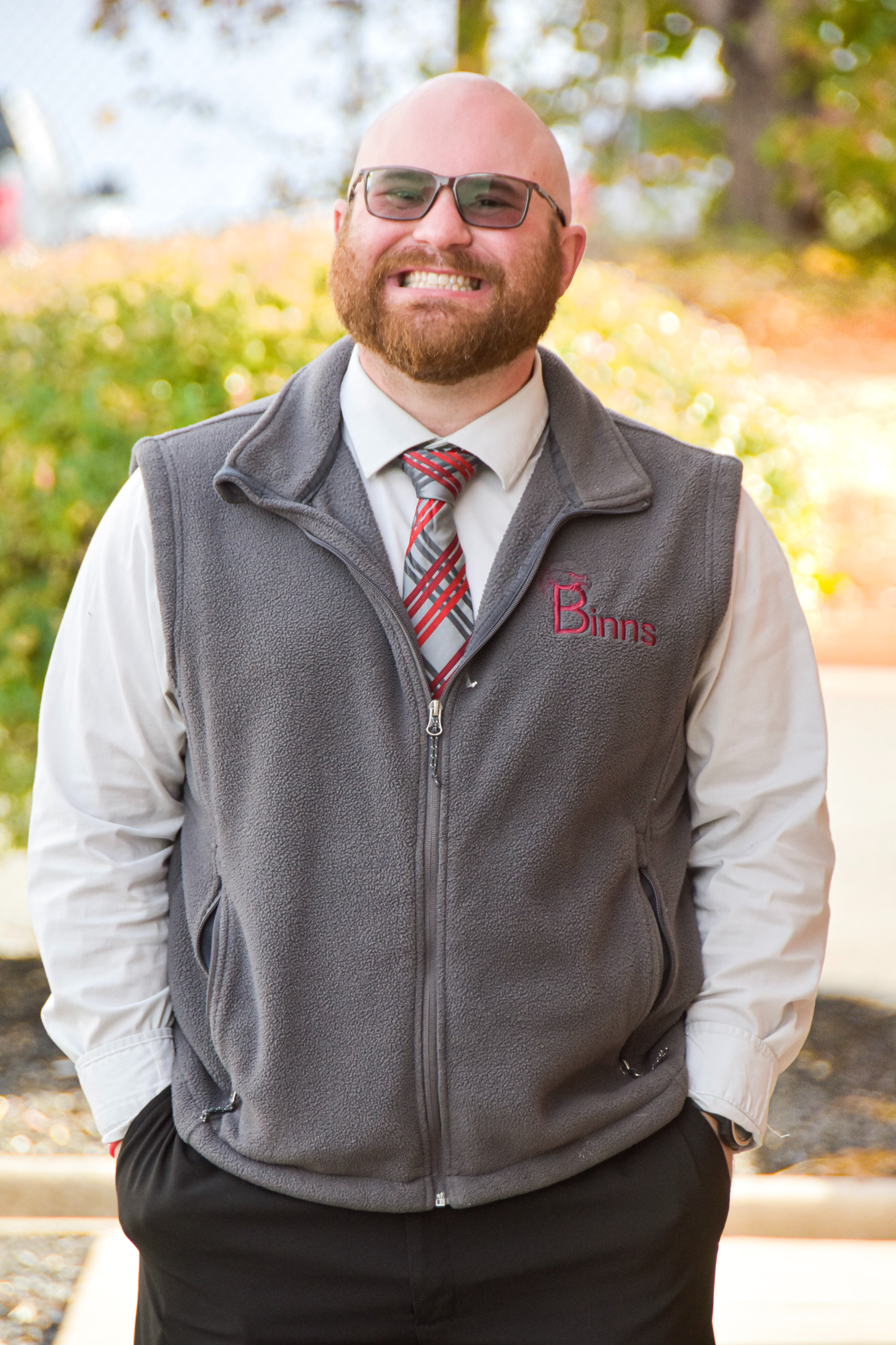 A man in a gray vest, plaid tie, and sunglasses stands outside with his hands in his pockets.