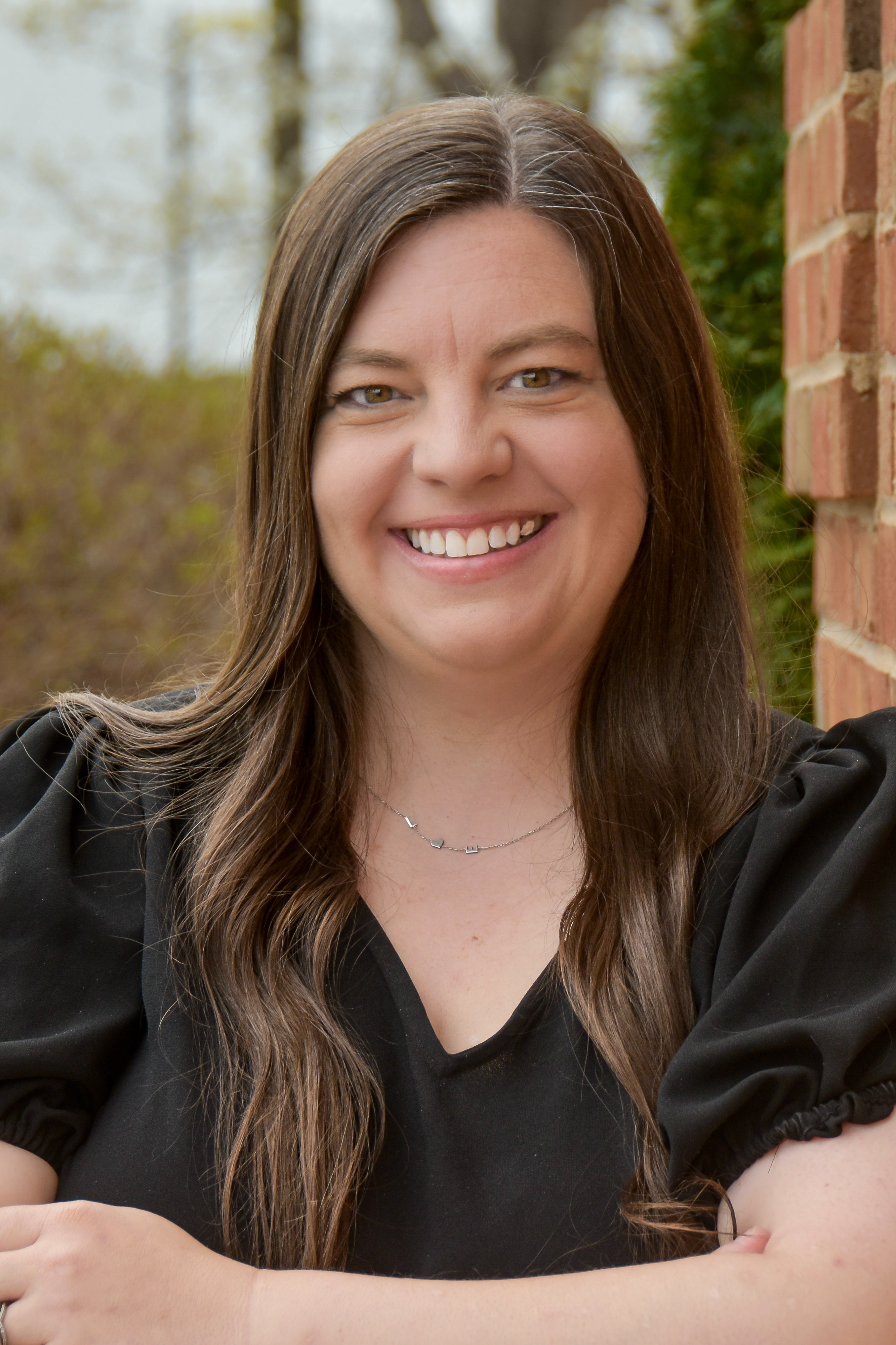 A smiling woman with long hair leans against a brick wall, wearing a black blouse with puffed sleeves.
