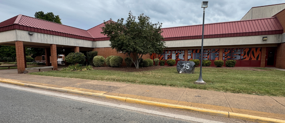 Building with red roof and white walls stands on street with lawn. Trees, bushes, and street light by curb.