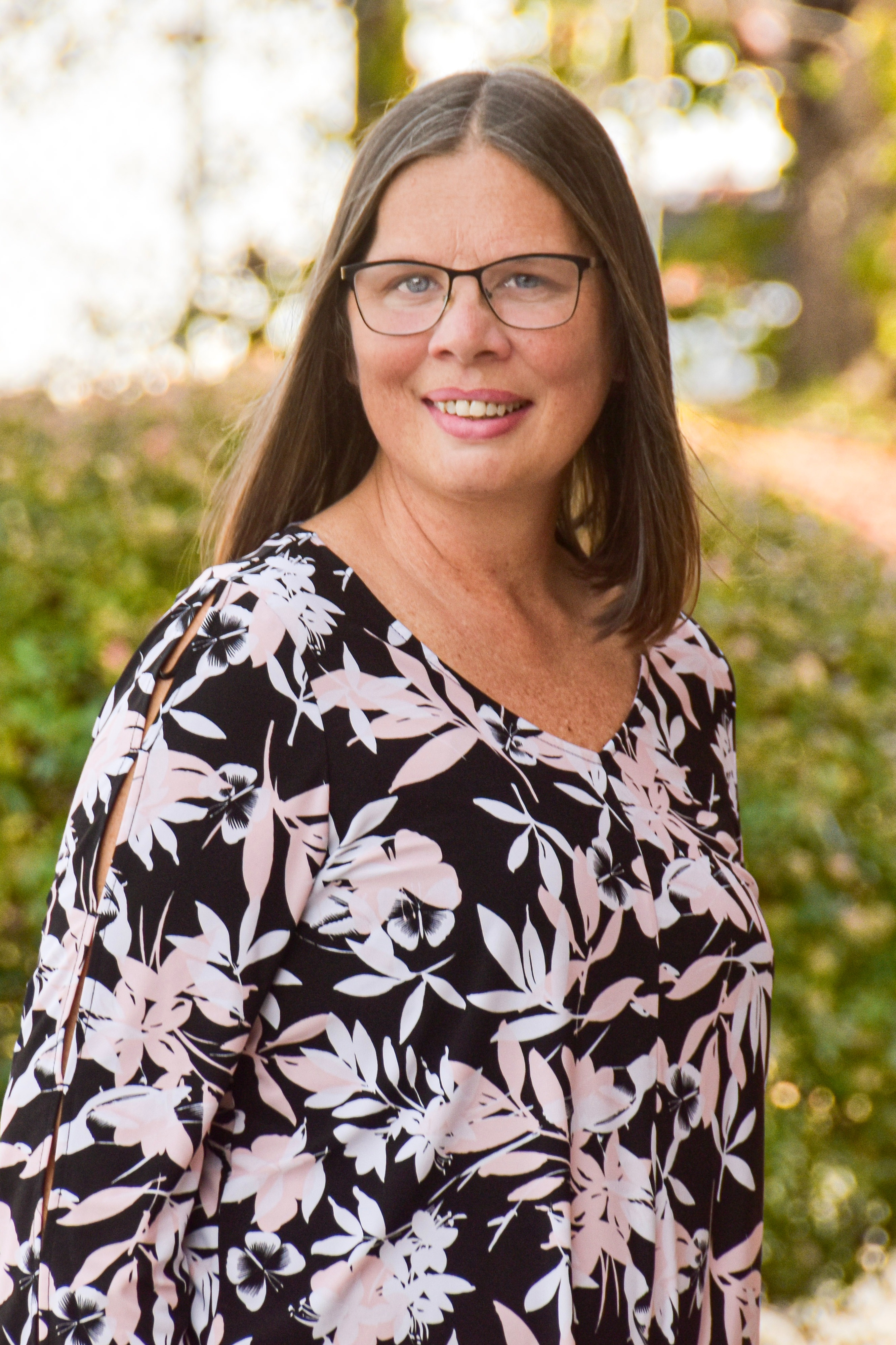Woman wearing glasses, black floral blouse, and carrying a white purse standing in front of green bushes.