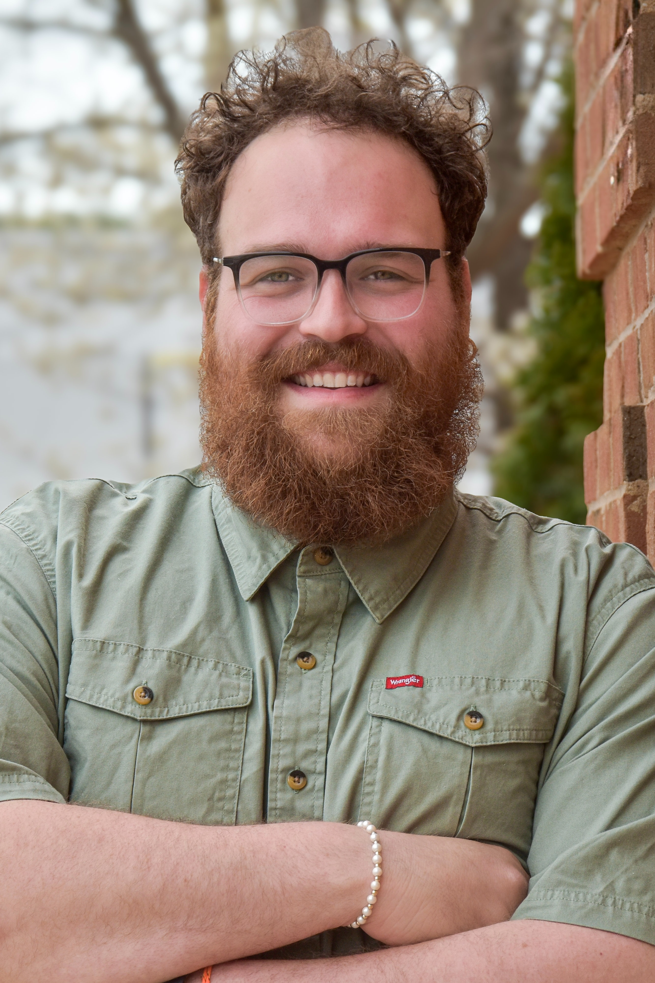 A man with a beard and glasses leans against a brick wall, wearing a green shirt. His arms are folded.