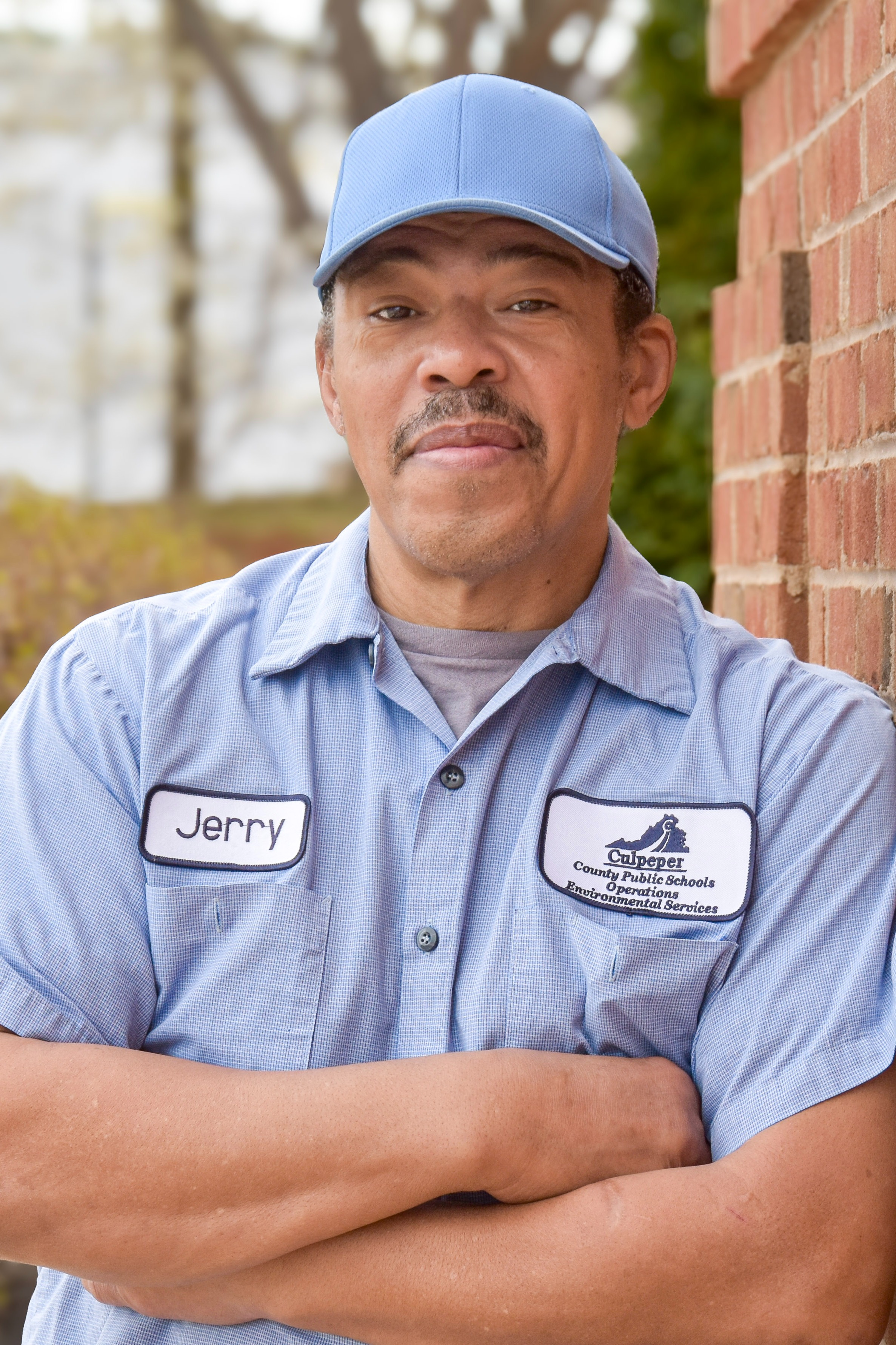 Man with arms crossed standing against a brick wall. He wears a blue shirt with a logo.