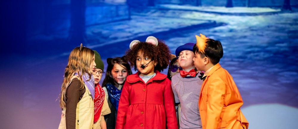 Children in costumes stand on a stage, one with a red coat and another with an orange outfit.